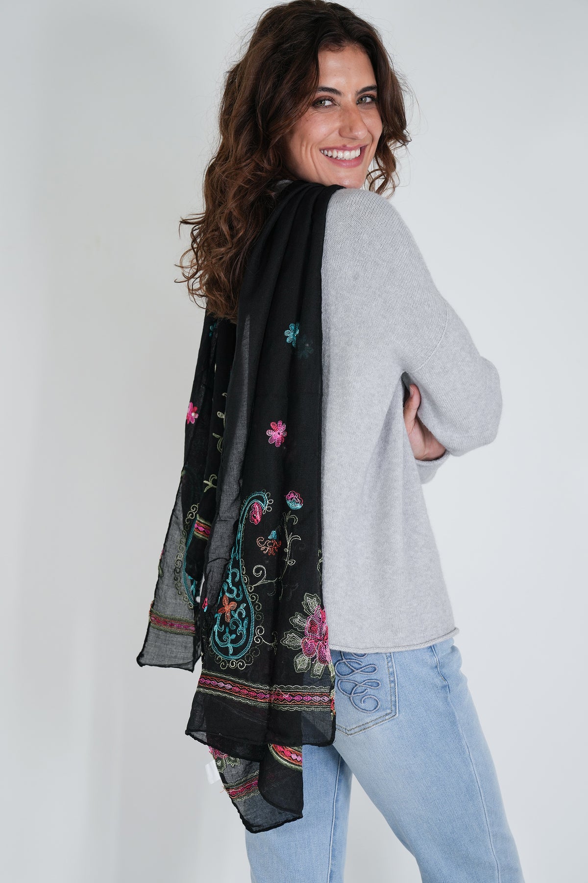 Woman wearing a black embroidered scarf, gray sweater, and light blue jeans on a white background