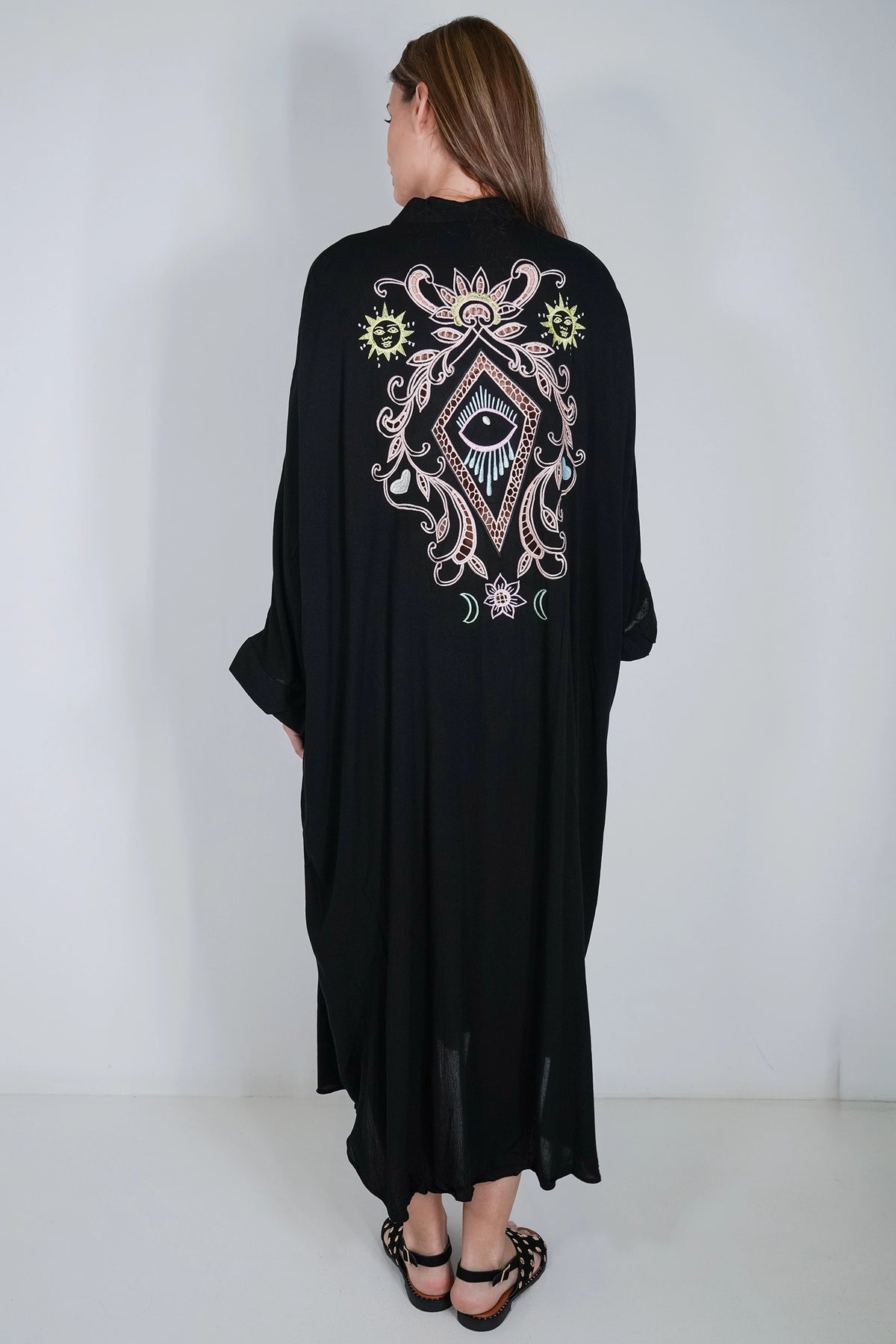 A woman viewed from the back wearing a long black kaftan-style dress with a large ornate embroidered design on the back featuring an eye motif and floral elements in pink, white, and green, paired with black strappy heels against a plain white studio background.