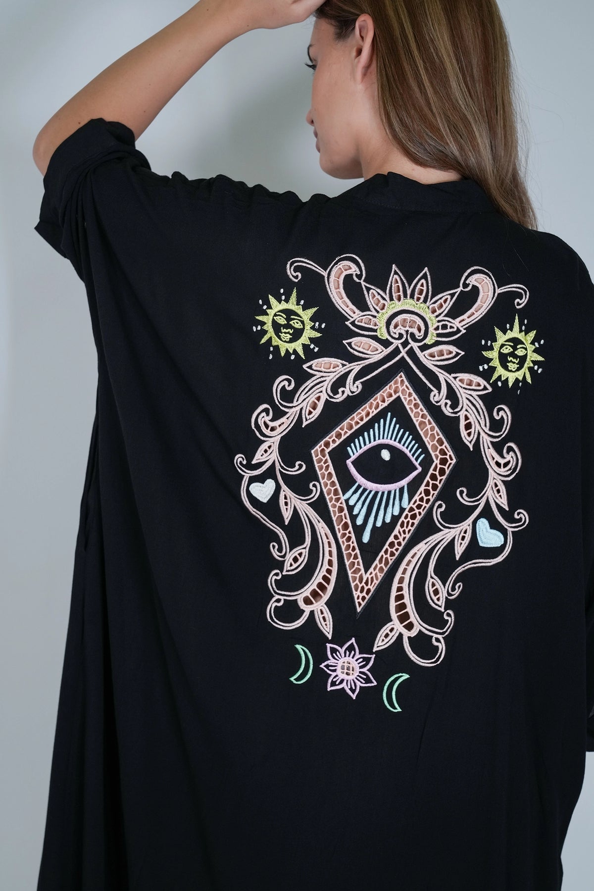 A woman wearing a black tunic-style top with a large multicolor graphic print on the back, featuring an eye, floral scrollwork, and sun motifs.