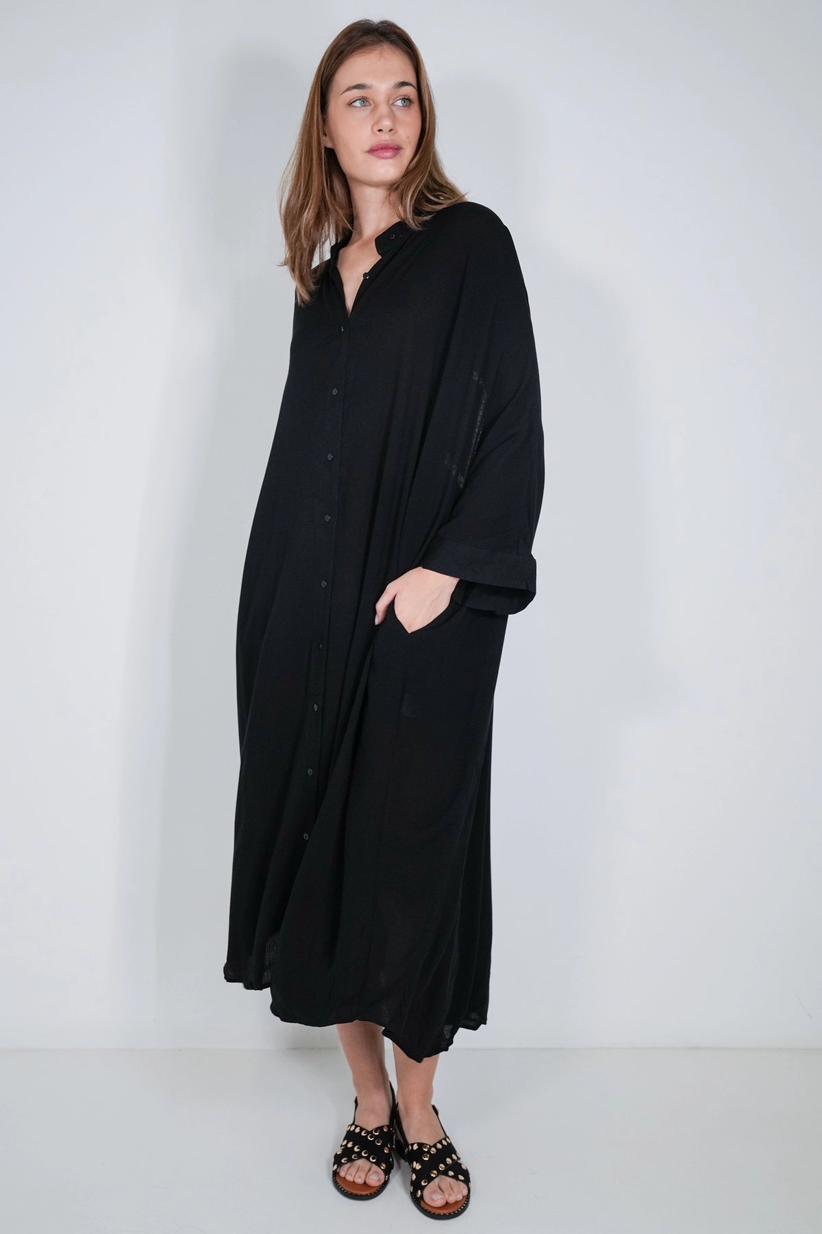 A woman modeling a long black button-front maxi dress with wide sleeves and side pockets, paired with black studded sandals, against a plain light gray studio background.