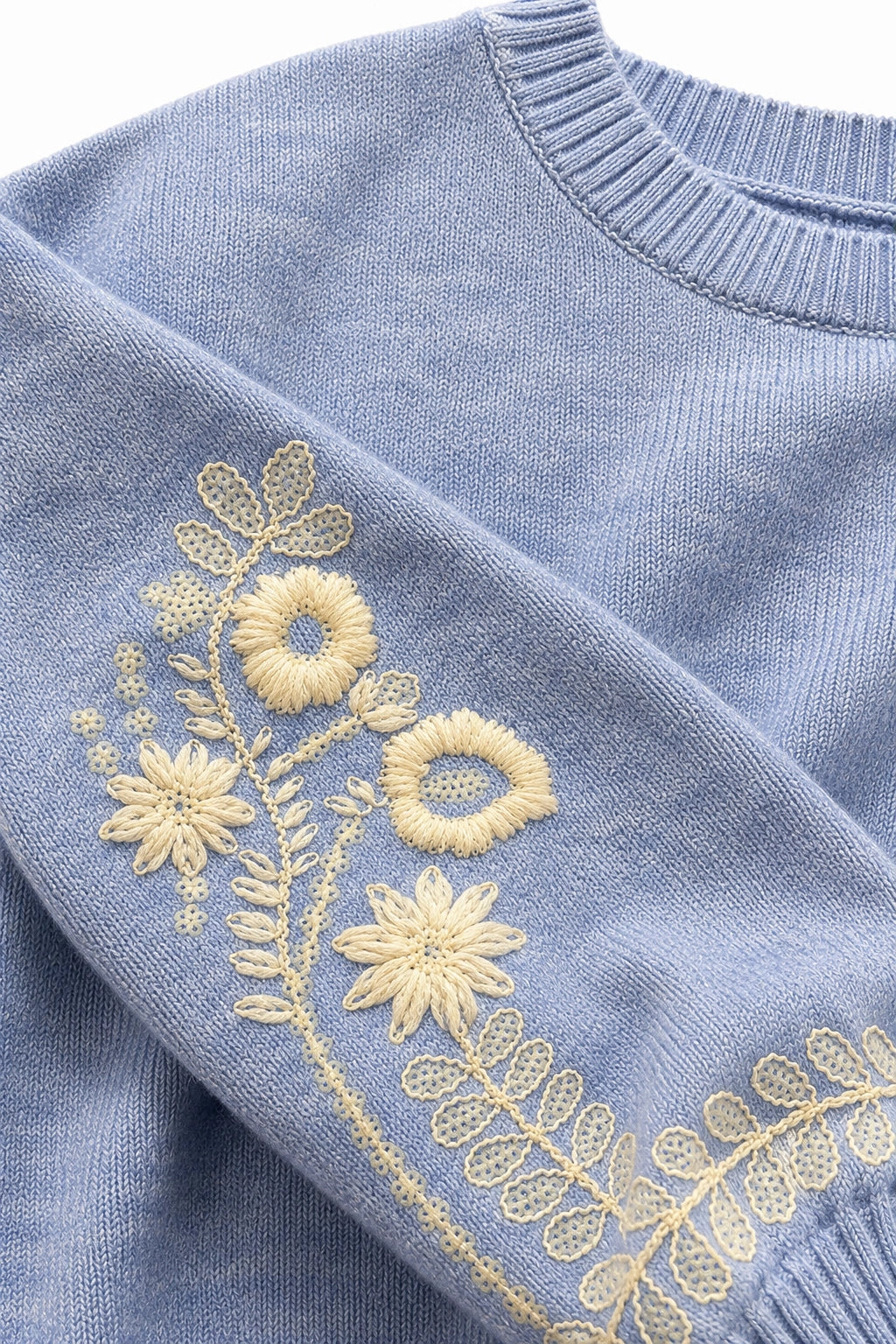 Close-up of a light blue knit sweater with cream-colored floral embroidery on the sleeve.