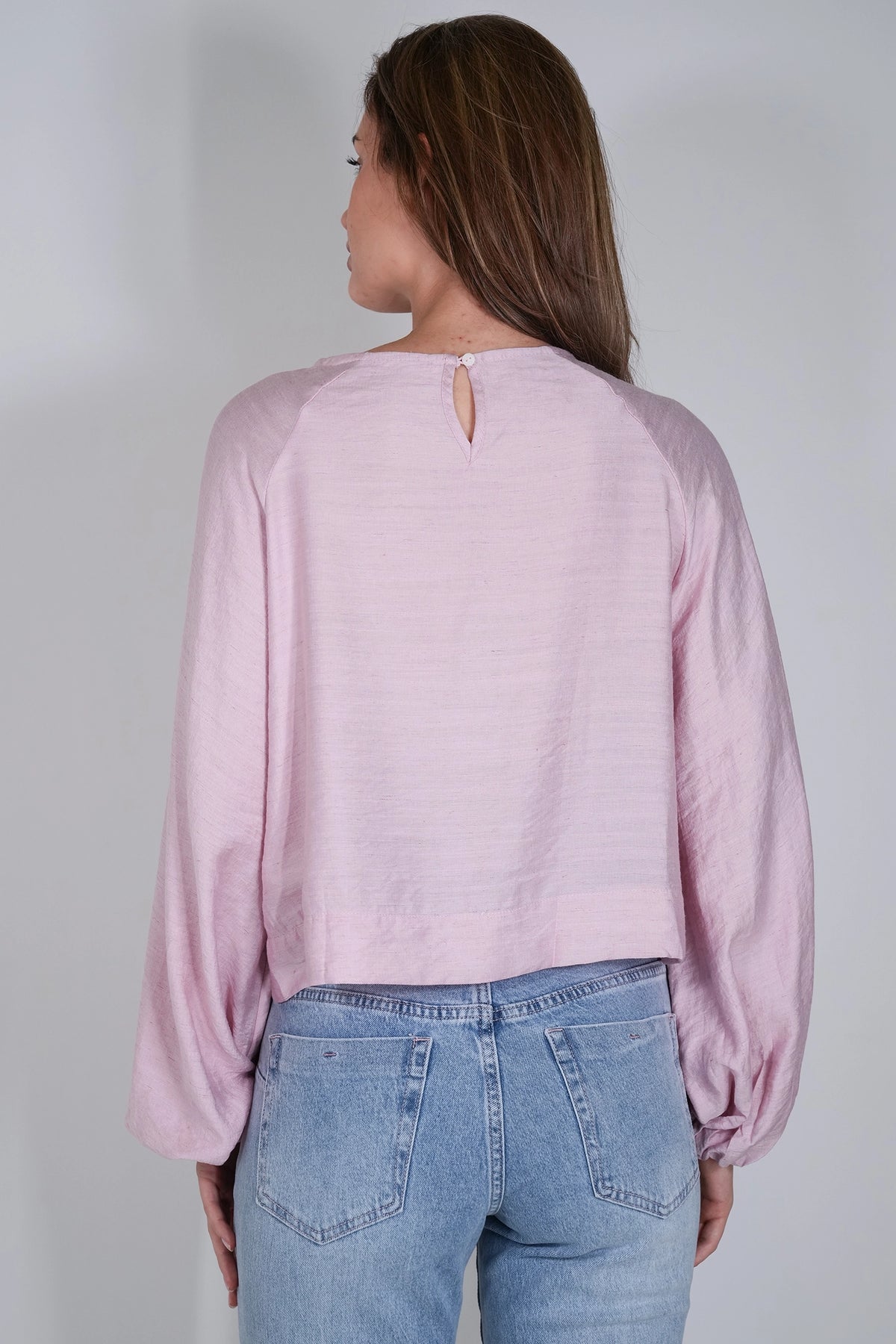 A woman shown from the back wearing a light pink long-sleeve top with a small keyhole cutout at the neckline, paired with light blue jeans, against a plain light gray background.