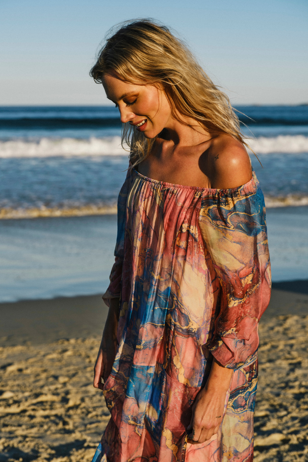 A woman standing on a sandy beach wearing a loose off-the-shoulder maxi dress with a colorful abstract print in pink, blue, and beige tones.