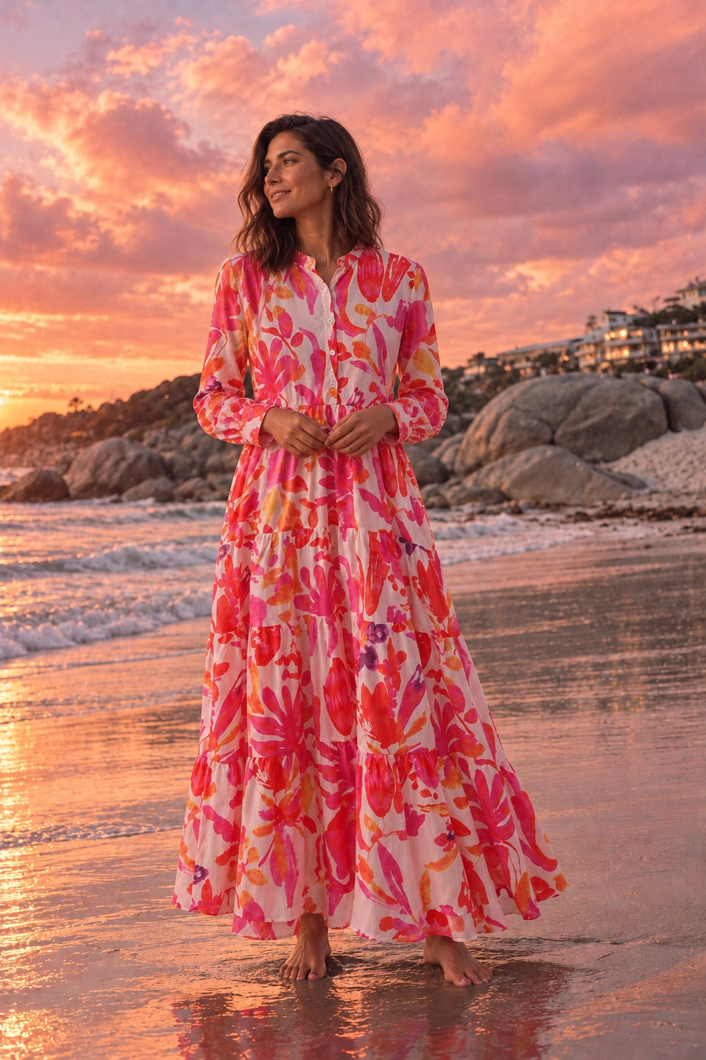 A woman stands barefoot on a beach at sunset wearing a long-sleeve, tiered maxi dress with a bold pink, red, orange, and white floral print.