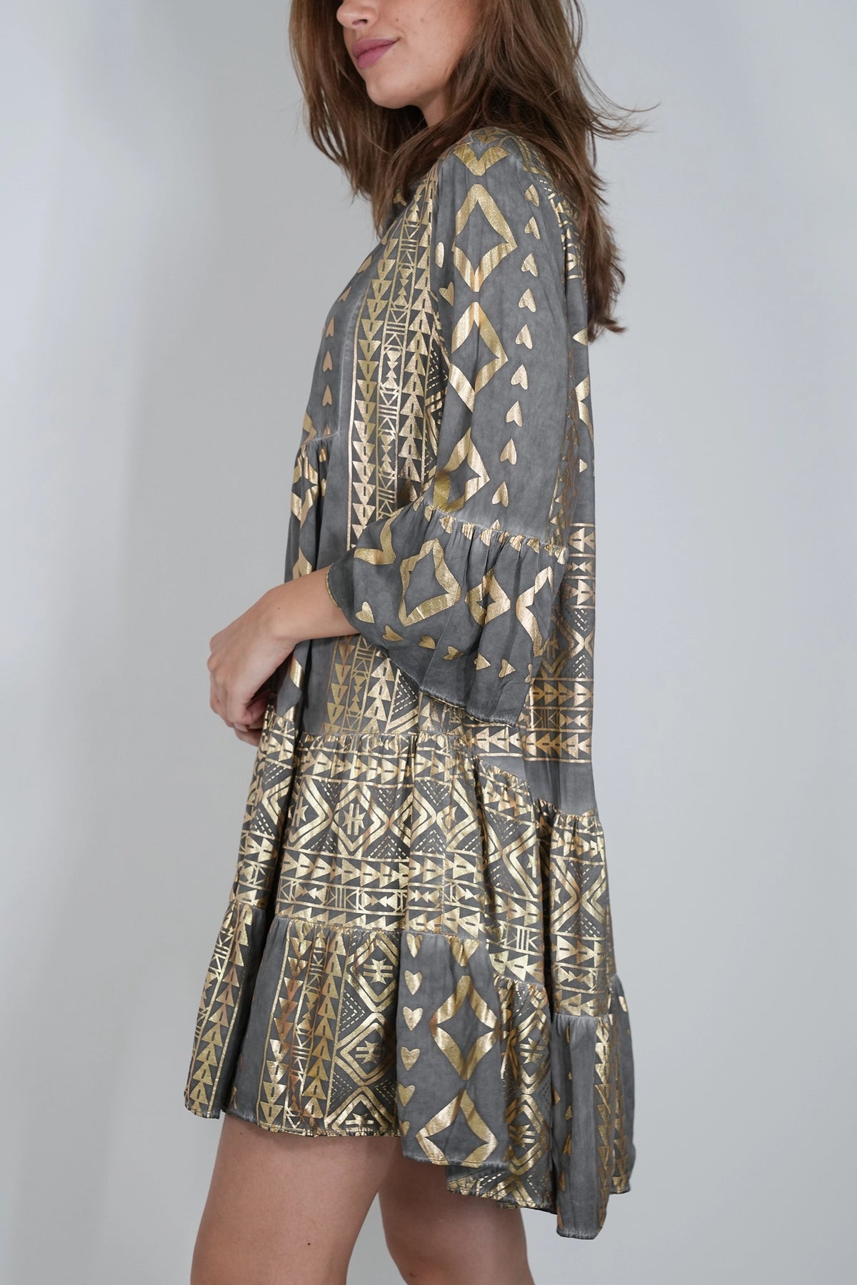 A woman wearing a gray and gold patterned long-sleeve mini dress, shown from a side-back angle against a light gray studio background.