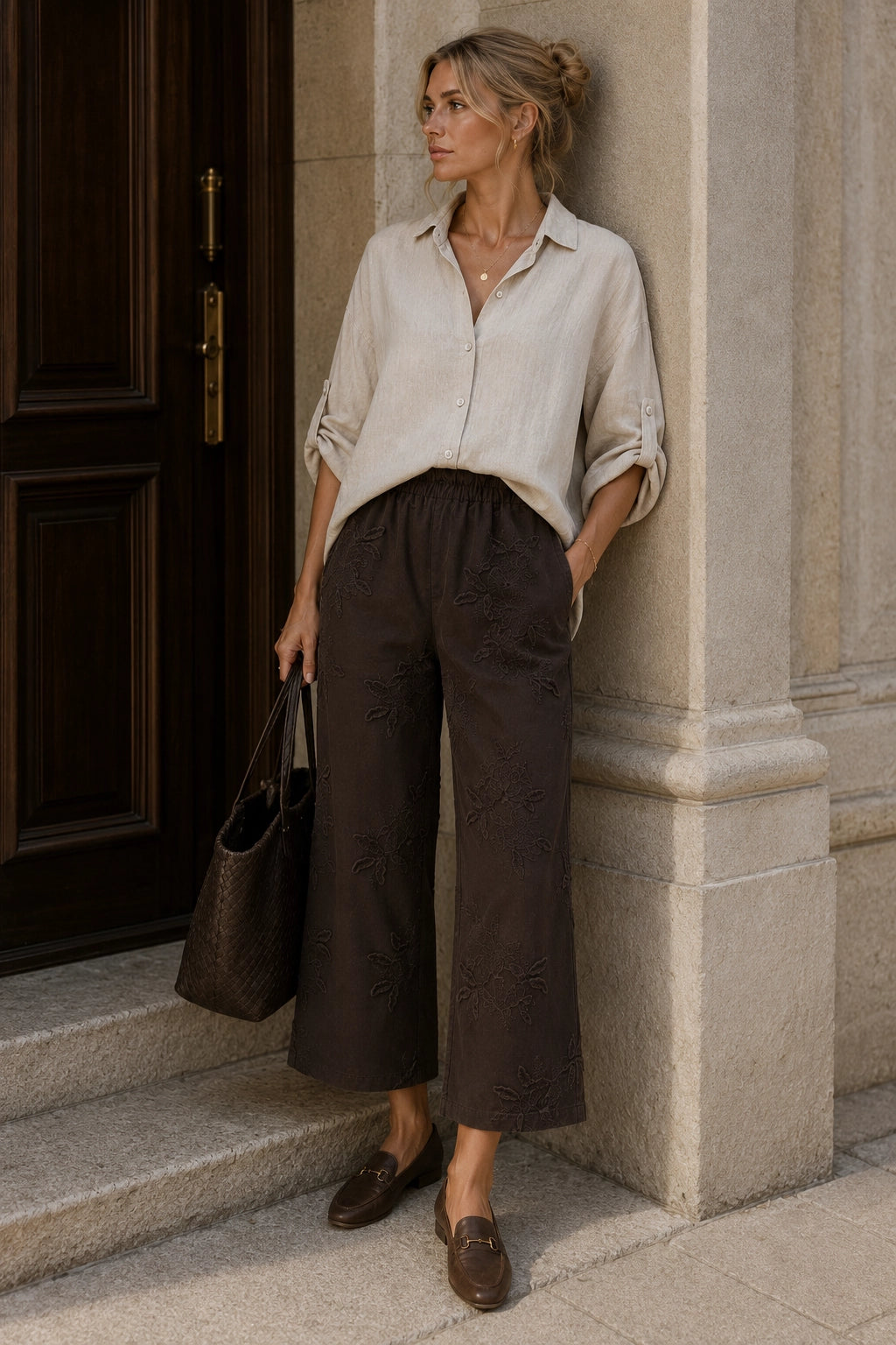 A woman stands on stone steps beside a dark wooden door, wearing a light beige button-up shirt, dark brown wide-leg cropped trousers, and brown loafers, while holding a dark brown tote bag.
