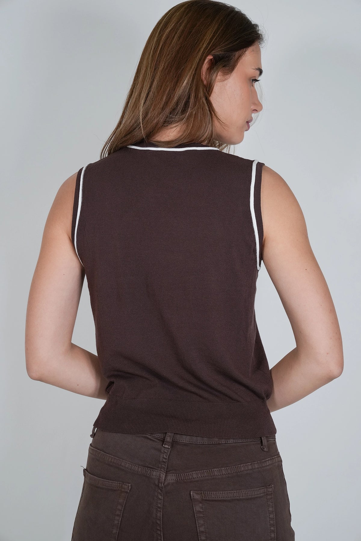 A woman shown from the back wearing a sleeveless dark brown top with white trim along the neckline and armholes, paired with dark brown pants, against a light gray studio background.