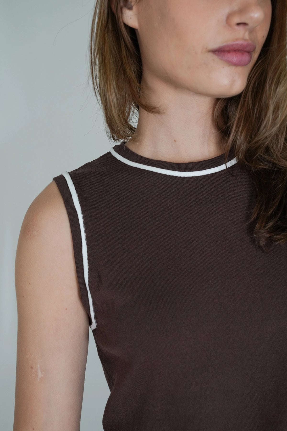 Close-up of a model wearing a dark brown sleeveless top with white contrast trim around the crew neck and armholes.