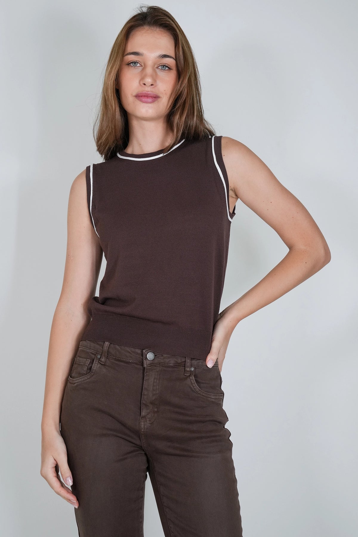 A woman modeling a sleeveless dark brown ribbed knit top with white contrast trim at the crew neck and armholes, paired with dark brown jeans, against a light gray studio background.