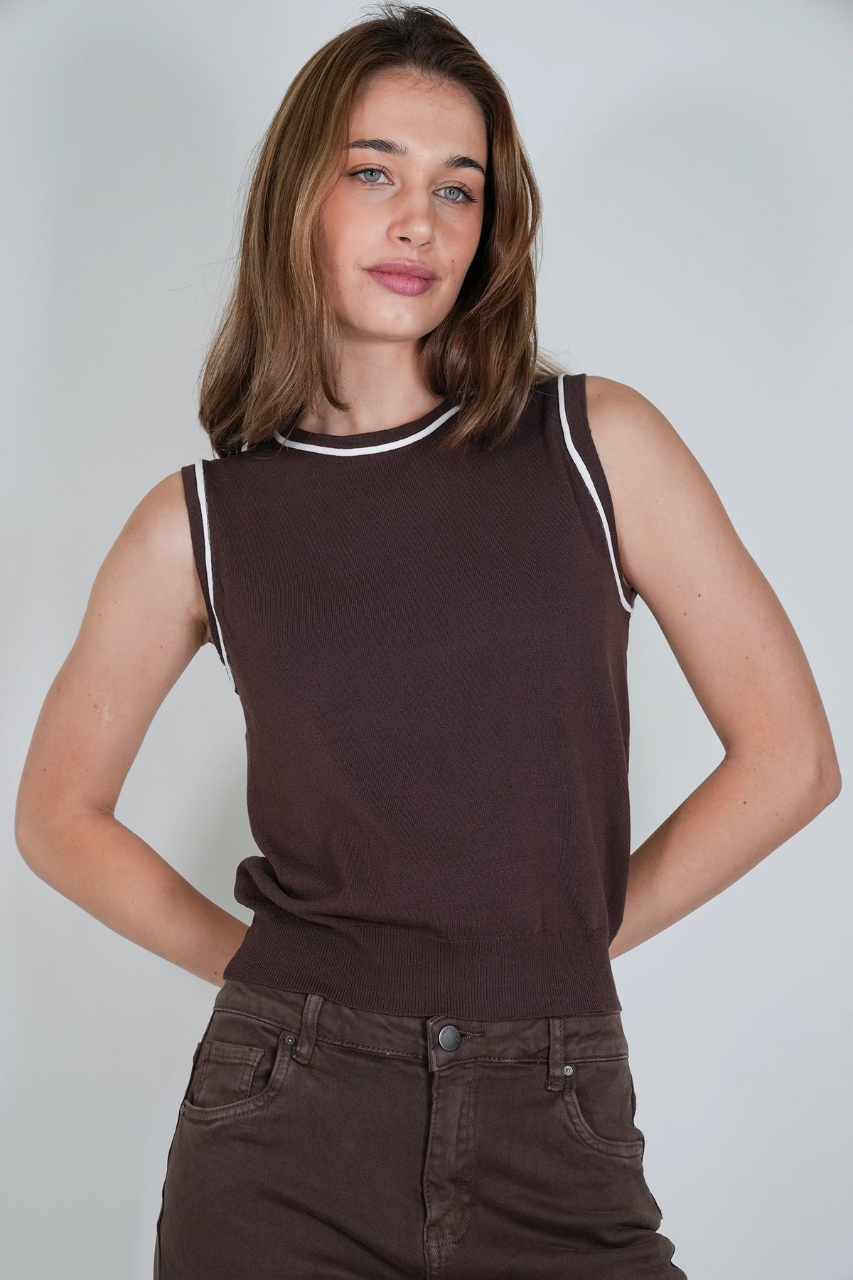 A woman wearing a sleeveless dark brown knit top with white contrast trim at the crew neck and armholes, paired with dark brown pants, against a light gray studio background.