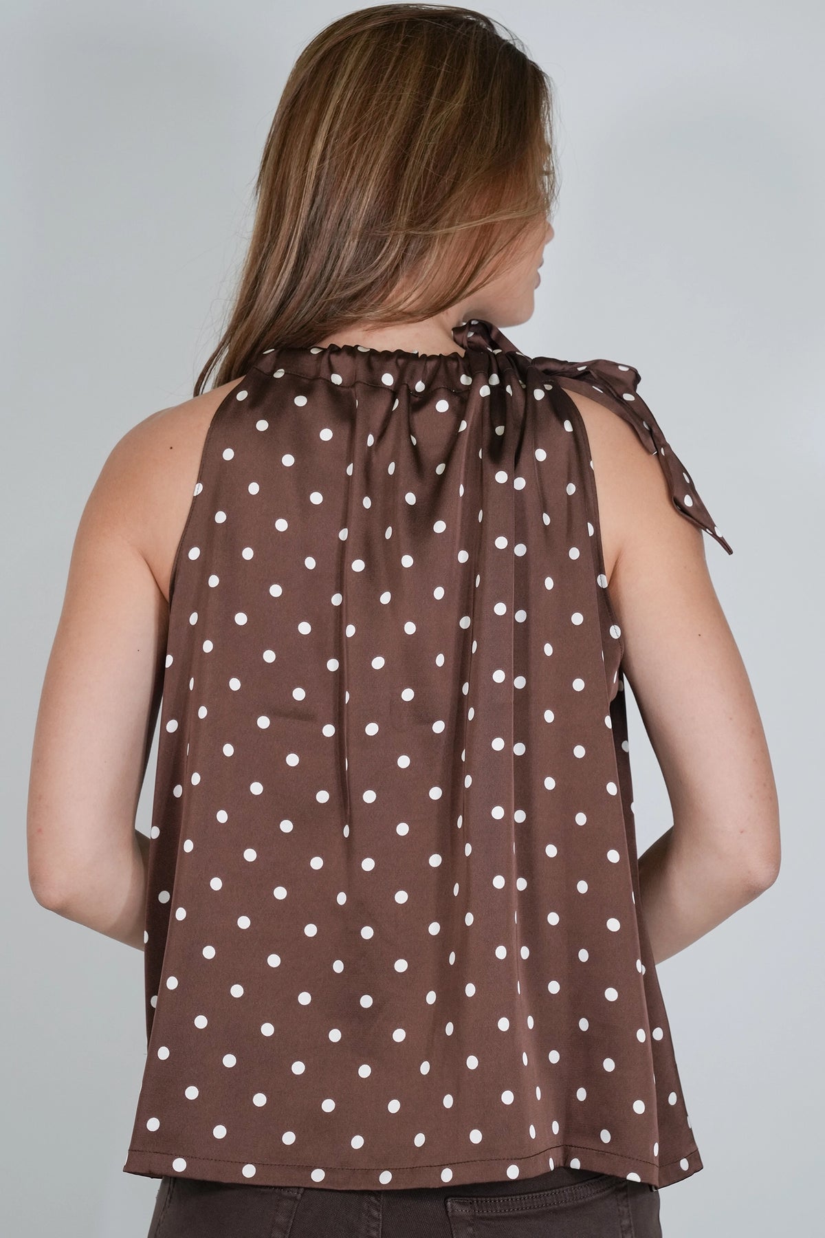 A woman shown from the back wearing a brown sleeveless top with a white polka dot pattern and a tie-neck detail, set against a plain light gray background.