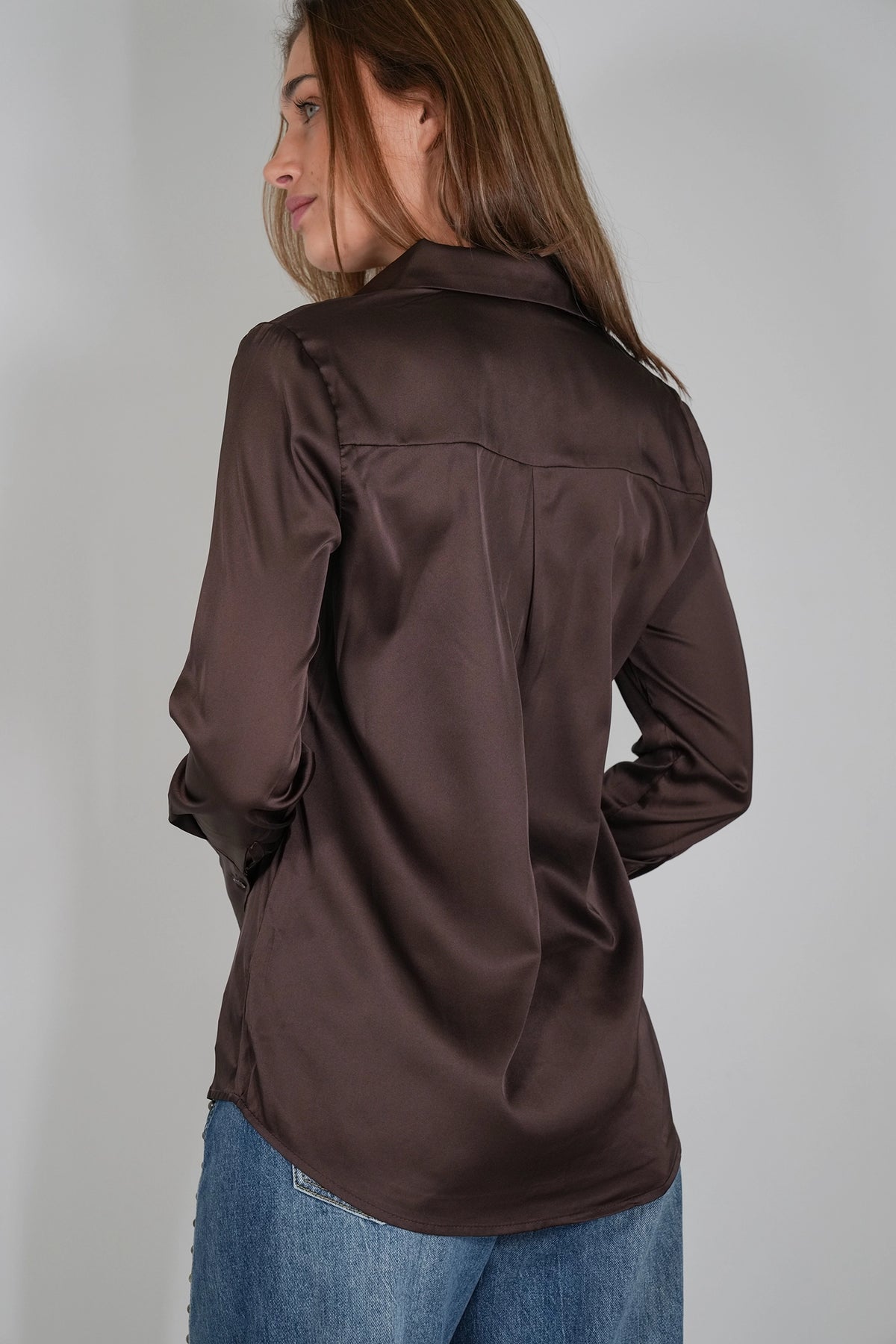 A woman wearing a dark brown satin long-sleeve button-up shirt, shown from a back-three-quarter angle against a plain light gray studio background.