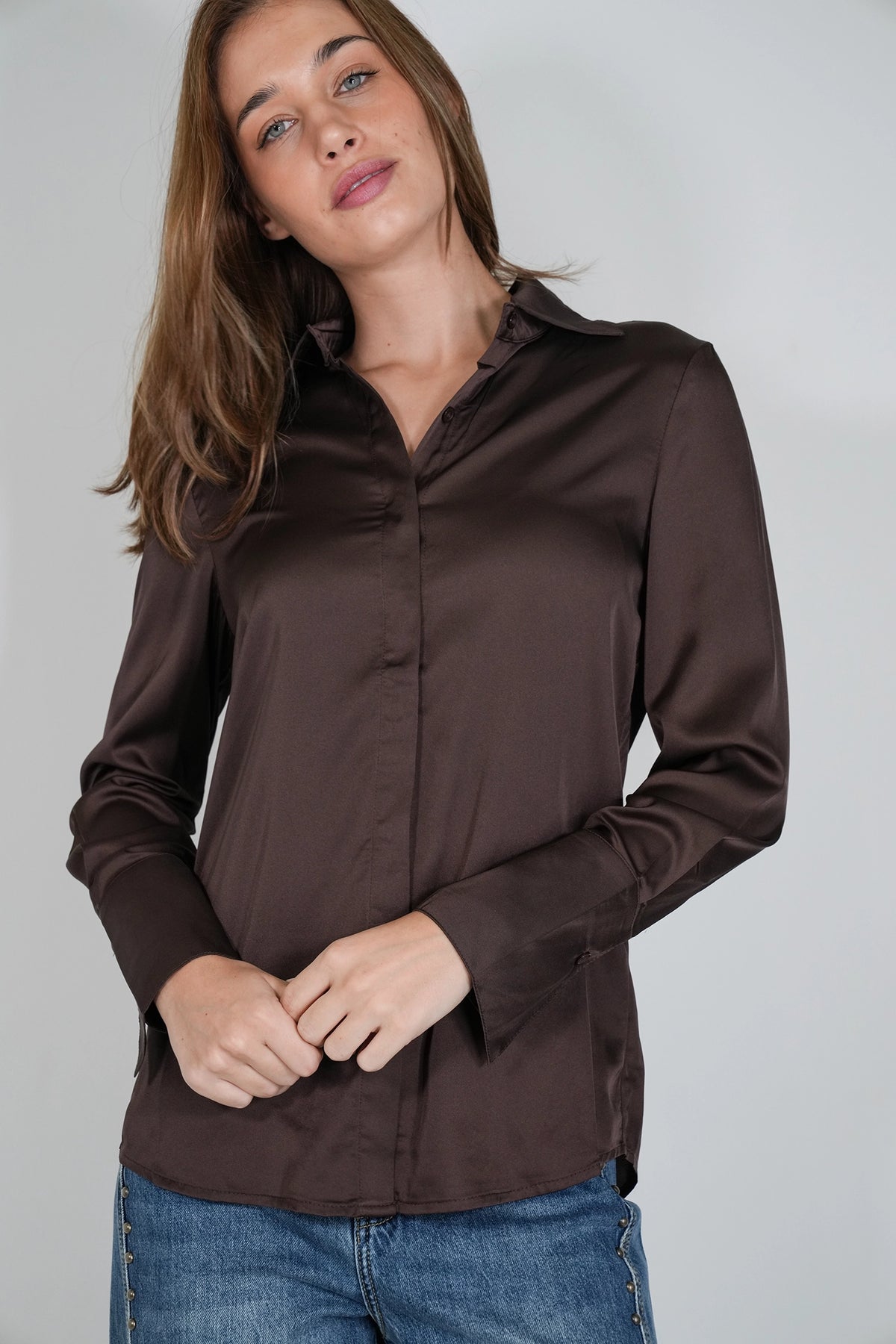 A woman wearing a dark brown long-sleeve satin button-up blouse and blue jeans, standing against a plain light gray studio background.