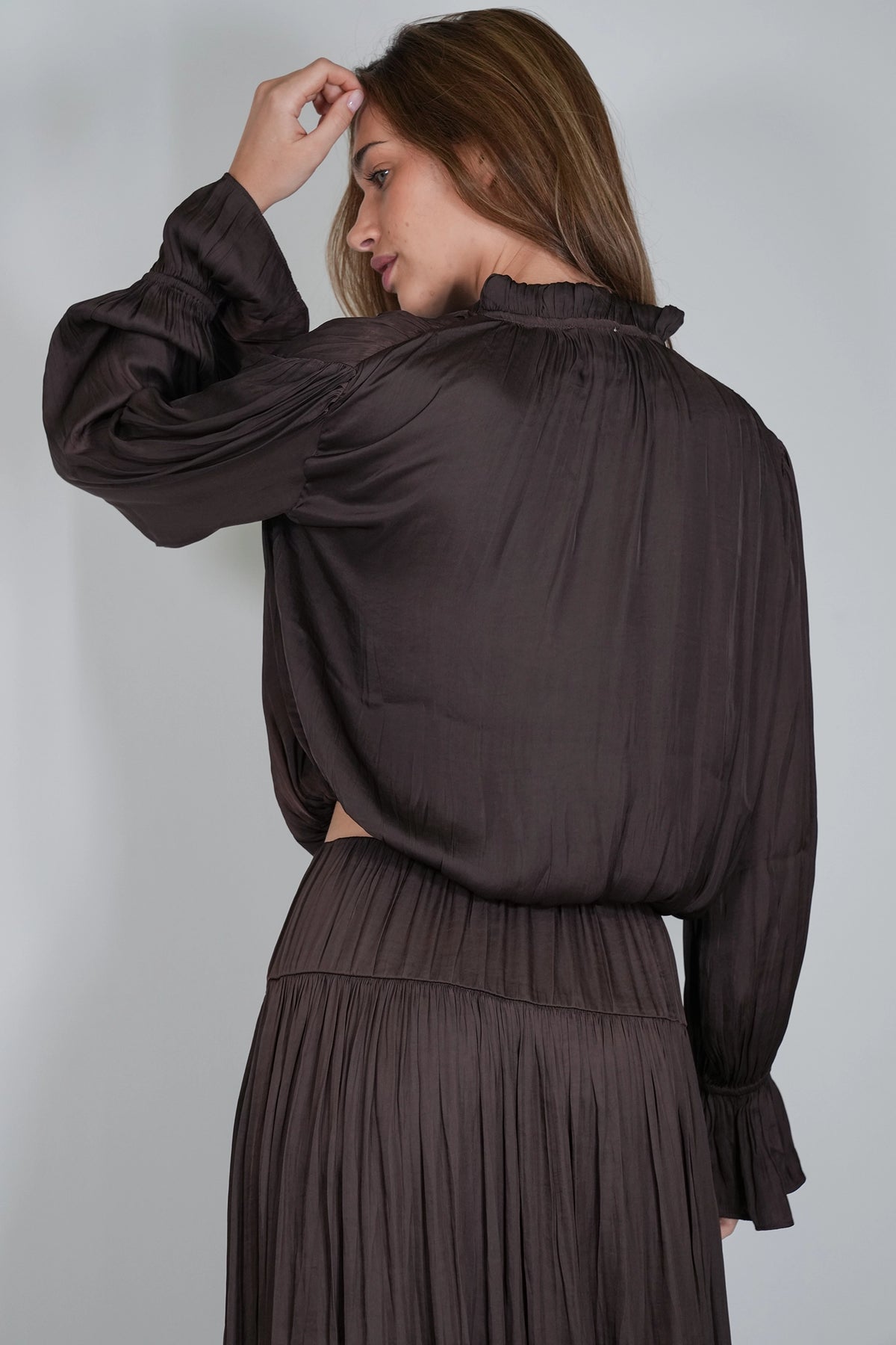 A woman wearing a dark brown long-sleeve blouse with a ruffled high neckline and a matching pleated skirt, shown from the back against a light gray studio background.