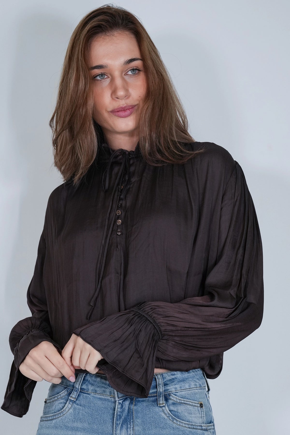 A woman wearing a dark brown long-sleeve blouse with a high neck, front buttons, and gathered ruffle cuffs, paired with blue jeans, against a light gray studio background.