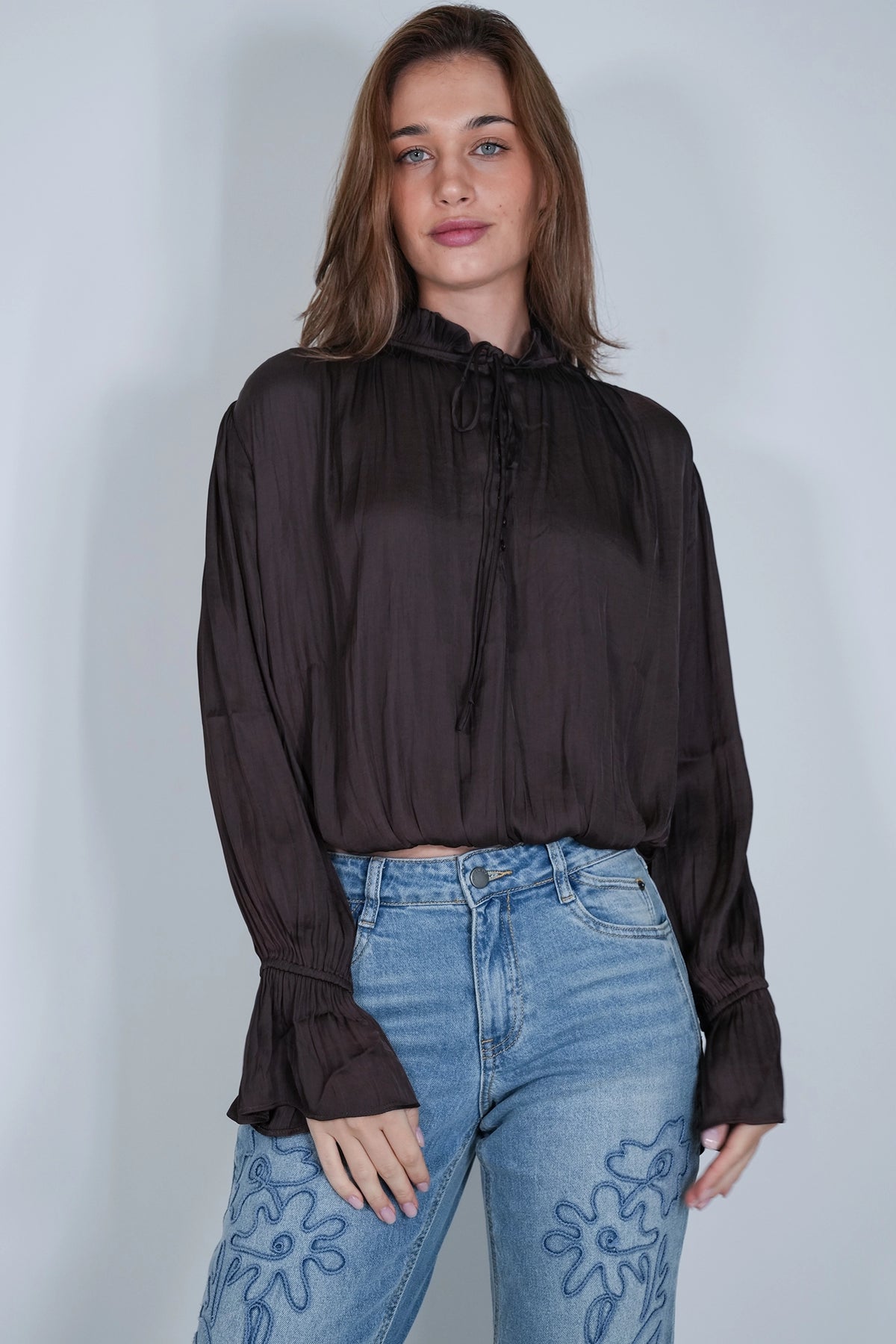 A woman wearing a dark brown sheer long-sleeve blouse with a high neck and drawstring, paired with light blue jeans featuring floral embroidery, standing against a light gray studio background.