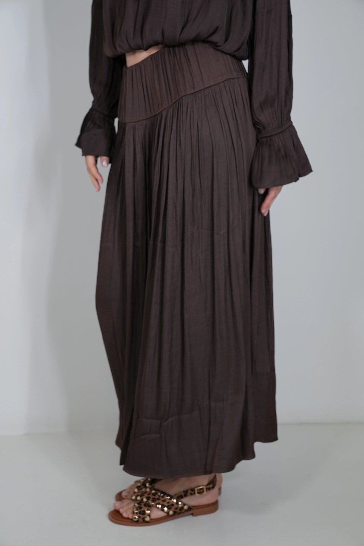 A person wearing a dark brown pleated maxi skirt with a matching long-sleeve top, paired with brown studded sandals, shown against a plain light gray studio background.