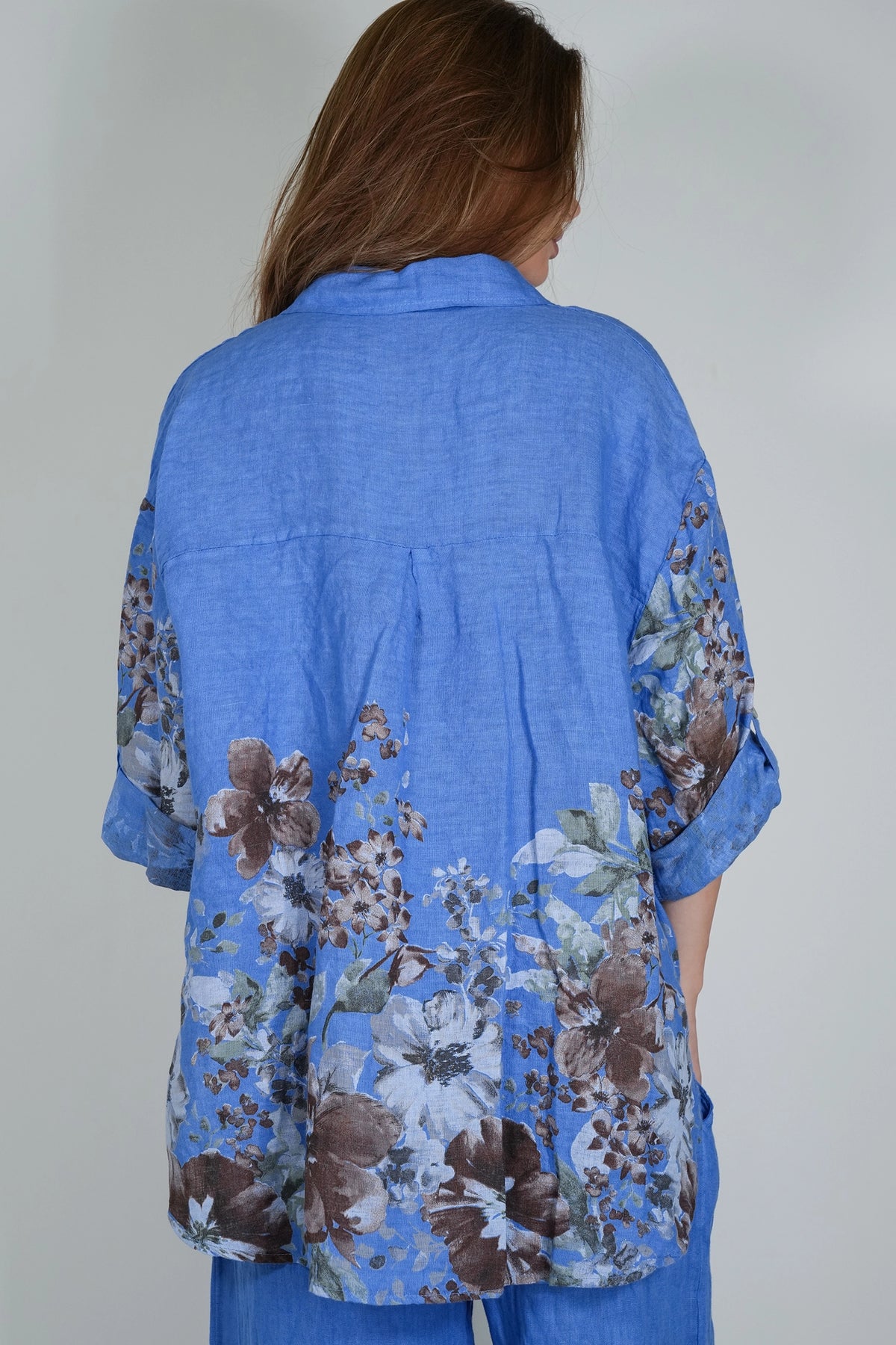 A woman viewed from behind wearing a blue short-sleeve linen shirt with a brown and white floral print on the lower back and sleeves, paired with blue pants against a light gray background.