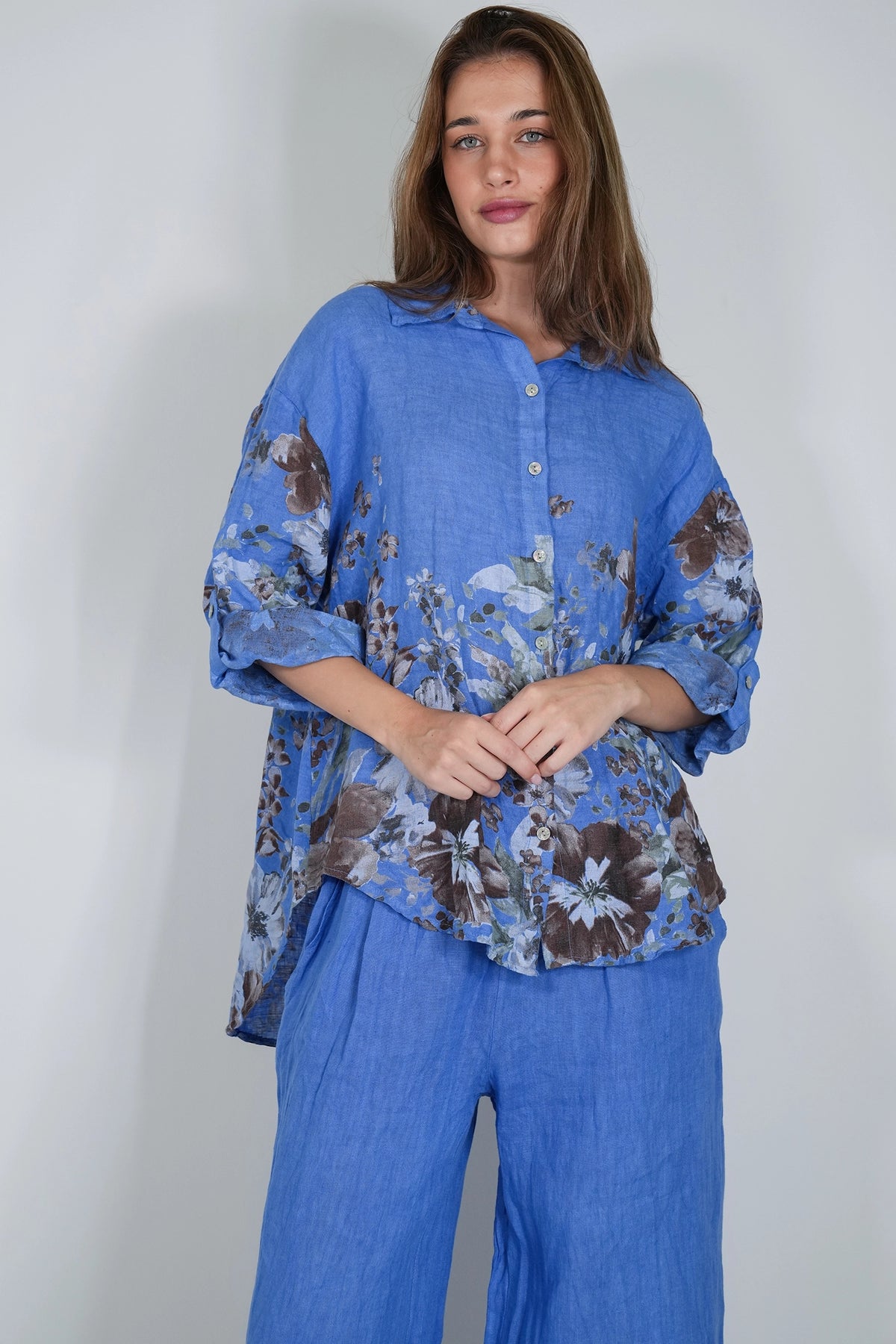 A woman modeling a blue button-up shirt with a brown and gray floral print, paired with matching blue pants, against a plain light gray background.