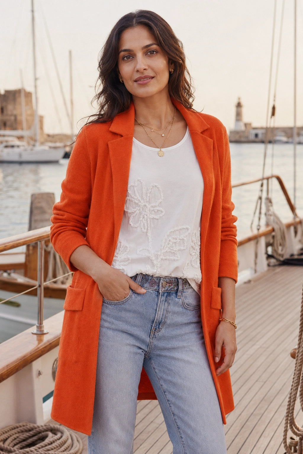 A woman stands on a wooden boat deck wearing a bright orange blazer over a white embroidered top and light blue jeans, with water and boats in the background.