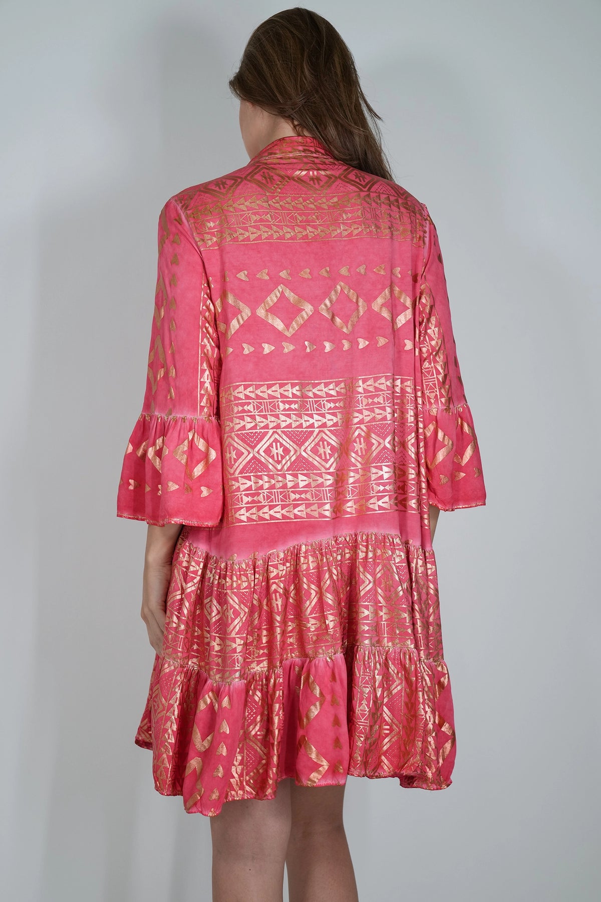 A woman viewed from the back wearing a pink bohemian-style tiered dress with gold geometric patterns, set against a plain light gray background.
