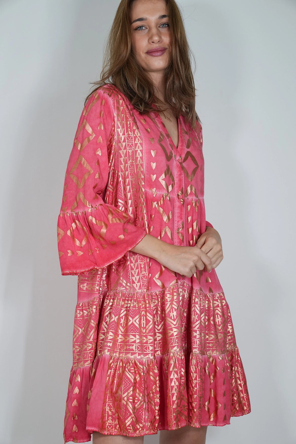 A woman wearing a pink tiered bohemian-style dress with gold geometric patterns, photographed against a plain light gray studio background.