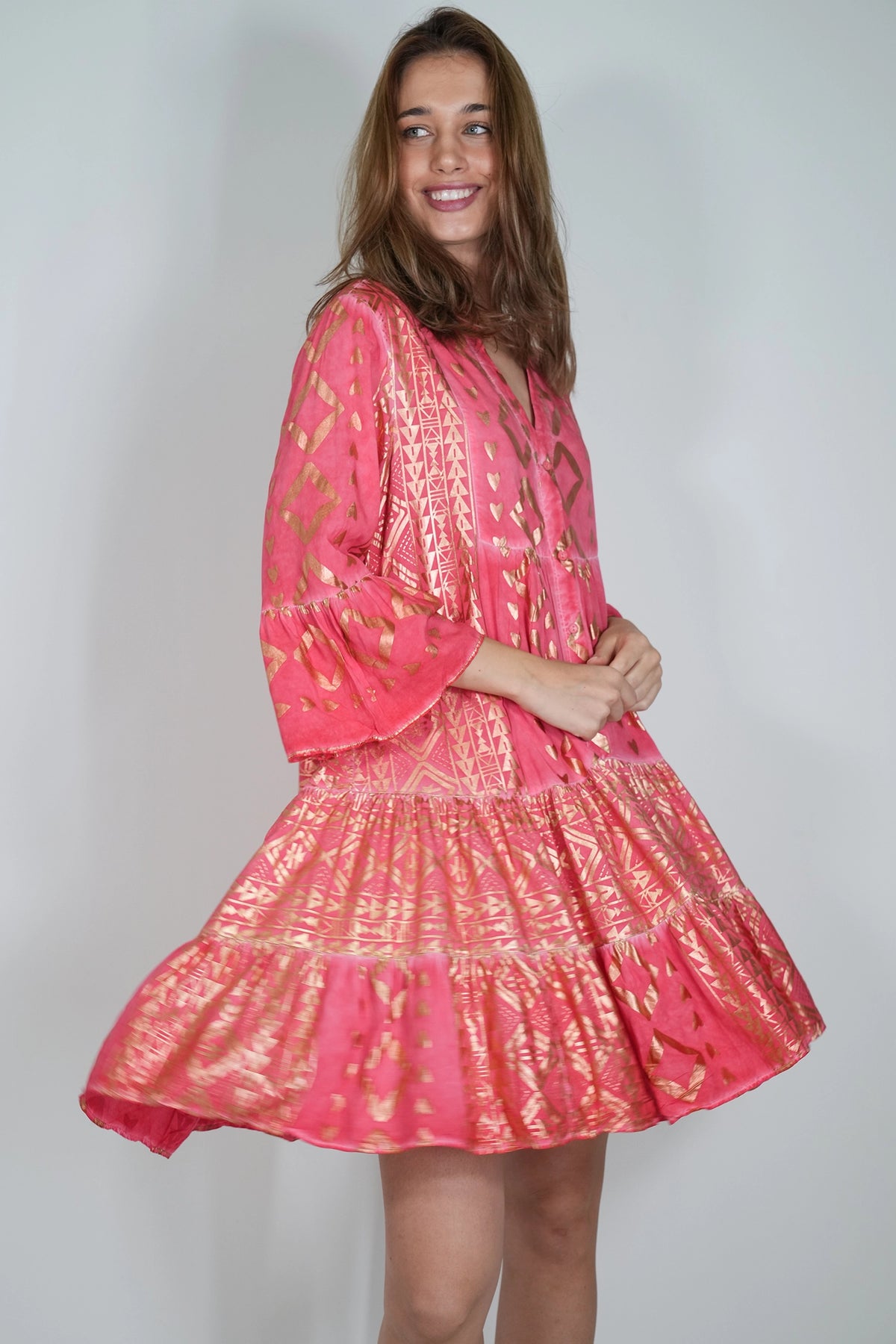 A woman modeling a bright pink tiered dress with gold metallic patterns, photographed against a plain light gray studio background.