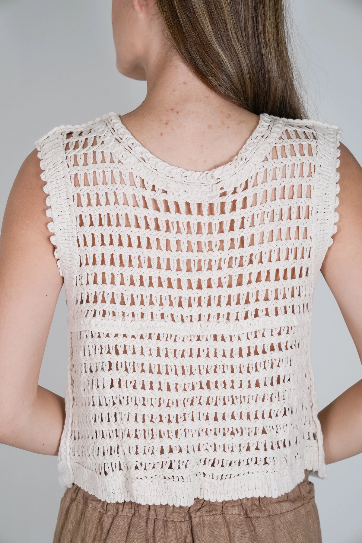 A woman shown from the back wearing a sleeveless crochet knit top in a warm off-white or cream color, paired with brown bottoms, against a plain light gray background.