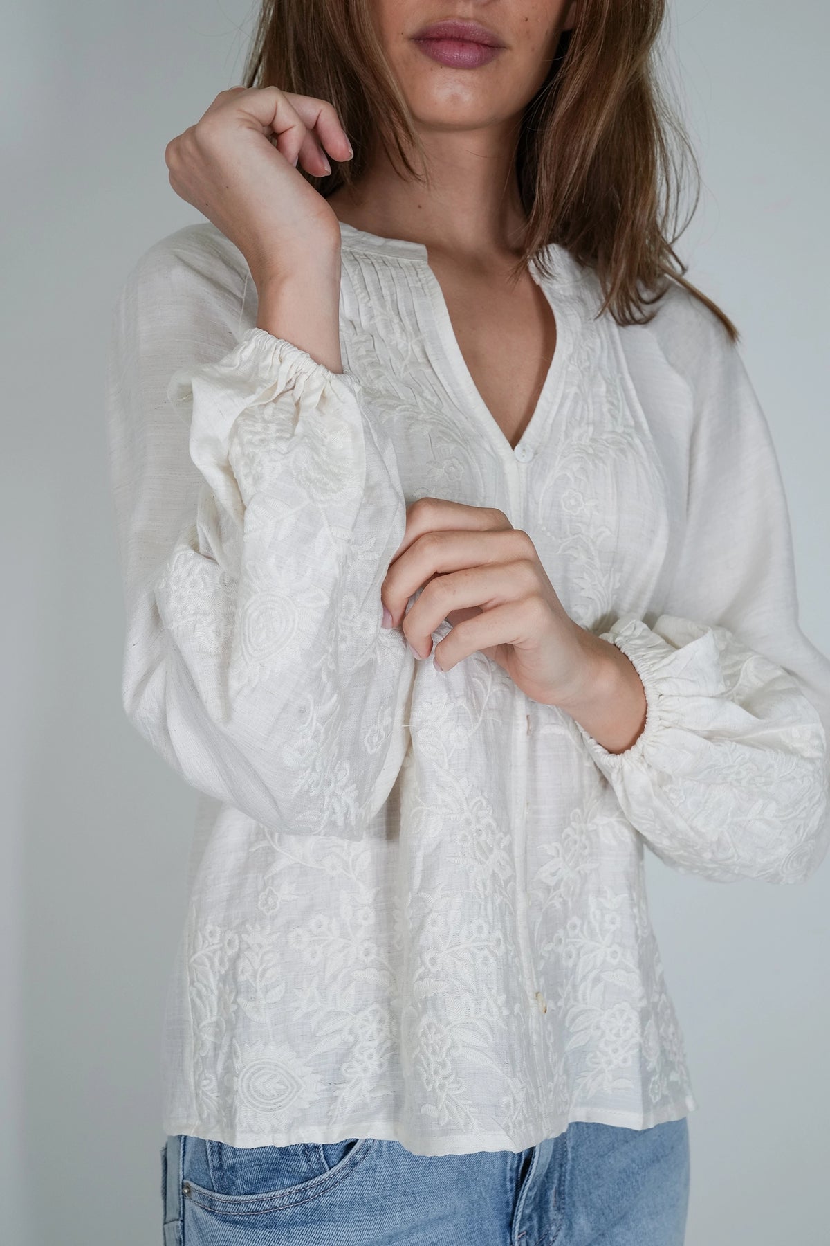 A woman wearing a white embroidered blouse with long balloon sleeves and a split neckline, paired with light blue jeans, photographed against a plain light gray background.