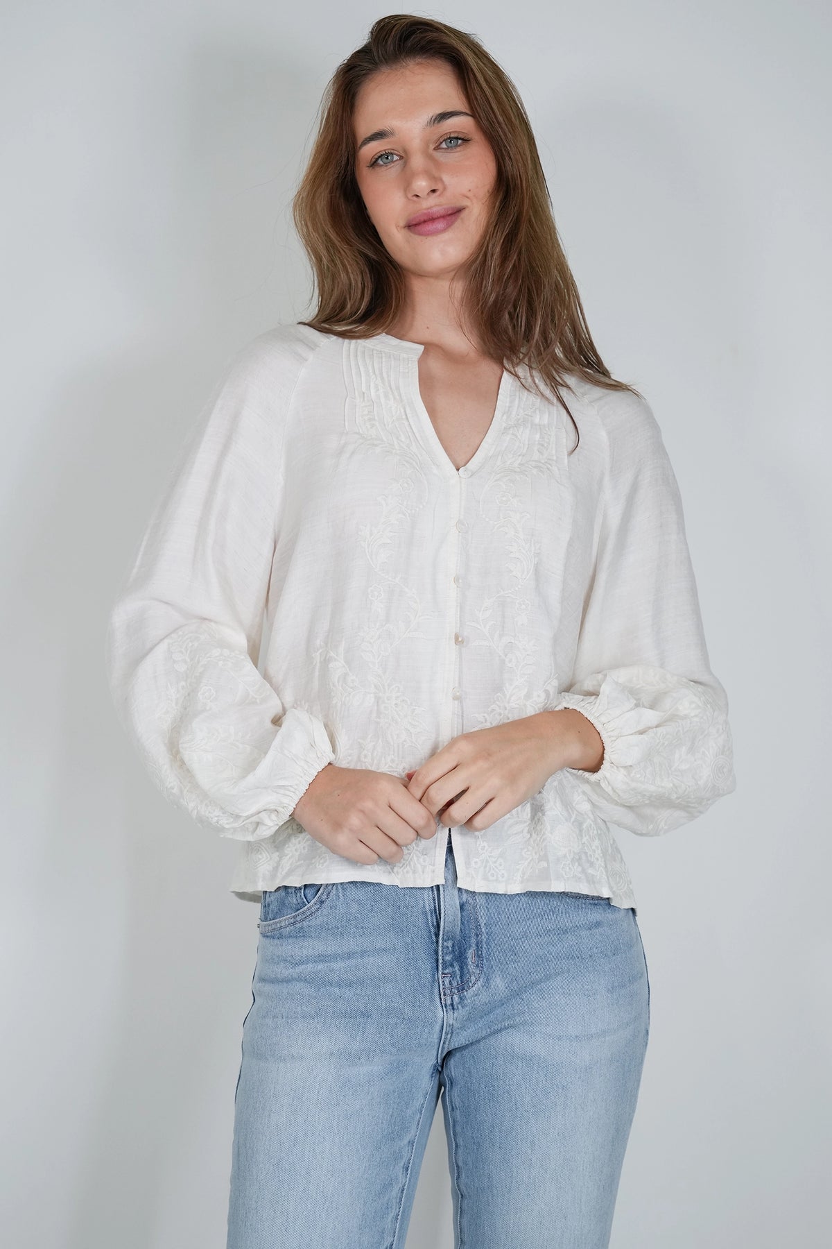 A woman wearing a white long-sleeve blouse with a split neckline and light blue jeans, standing against a plain light gray studio background.