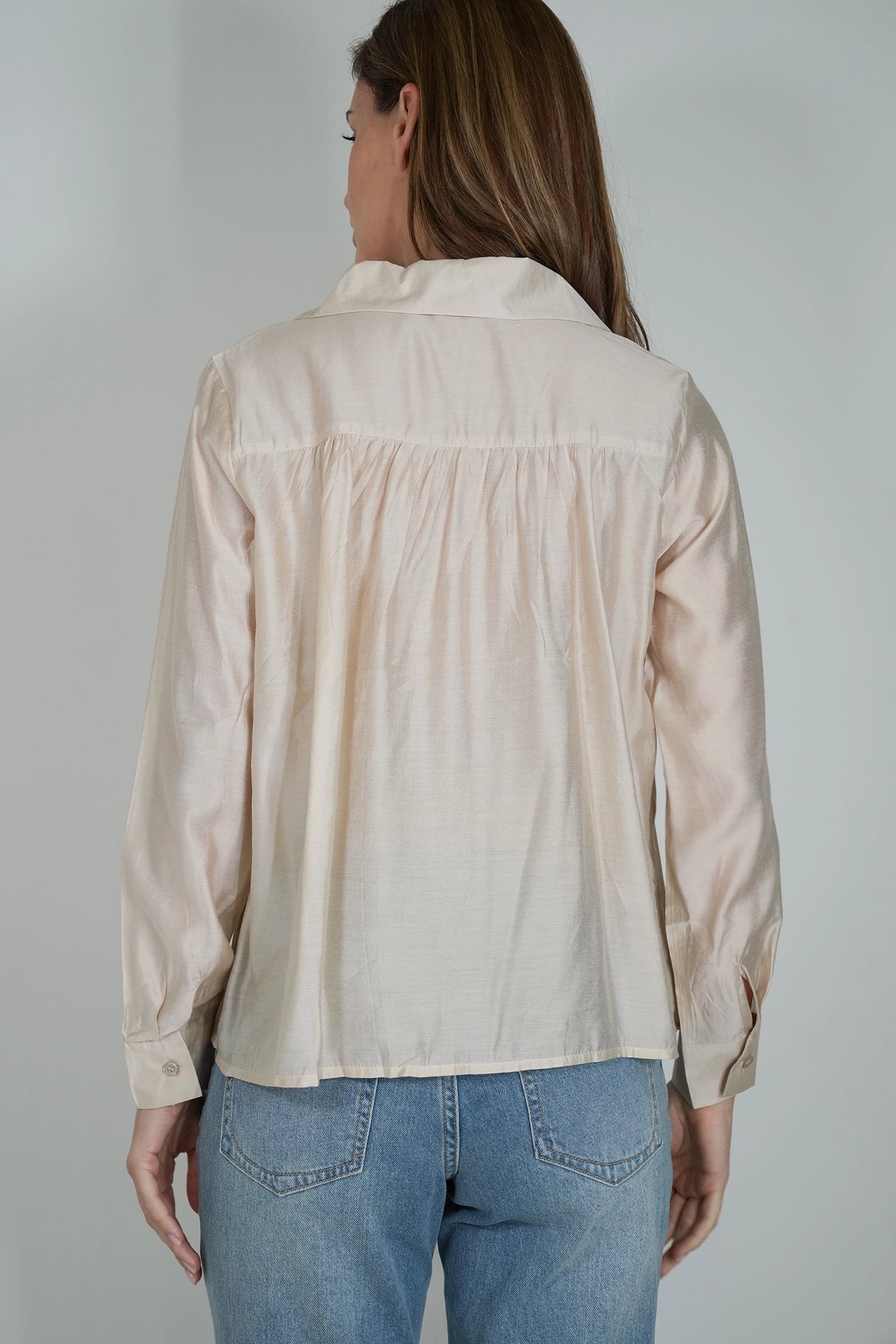 A woman viewed from the back wearing a light beige long-sleeve blouse with a gathered back detail and blue jeans, set against a plain light gray studio background.