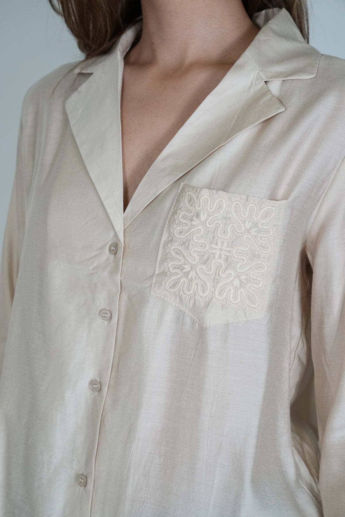 A close-up of a person wearing a light cream or off-white button-front shirt with a pointed collar and a decorative embroidered chest pocket.