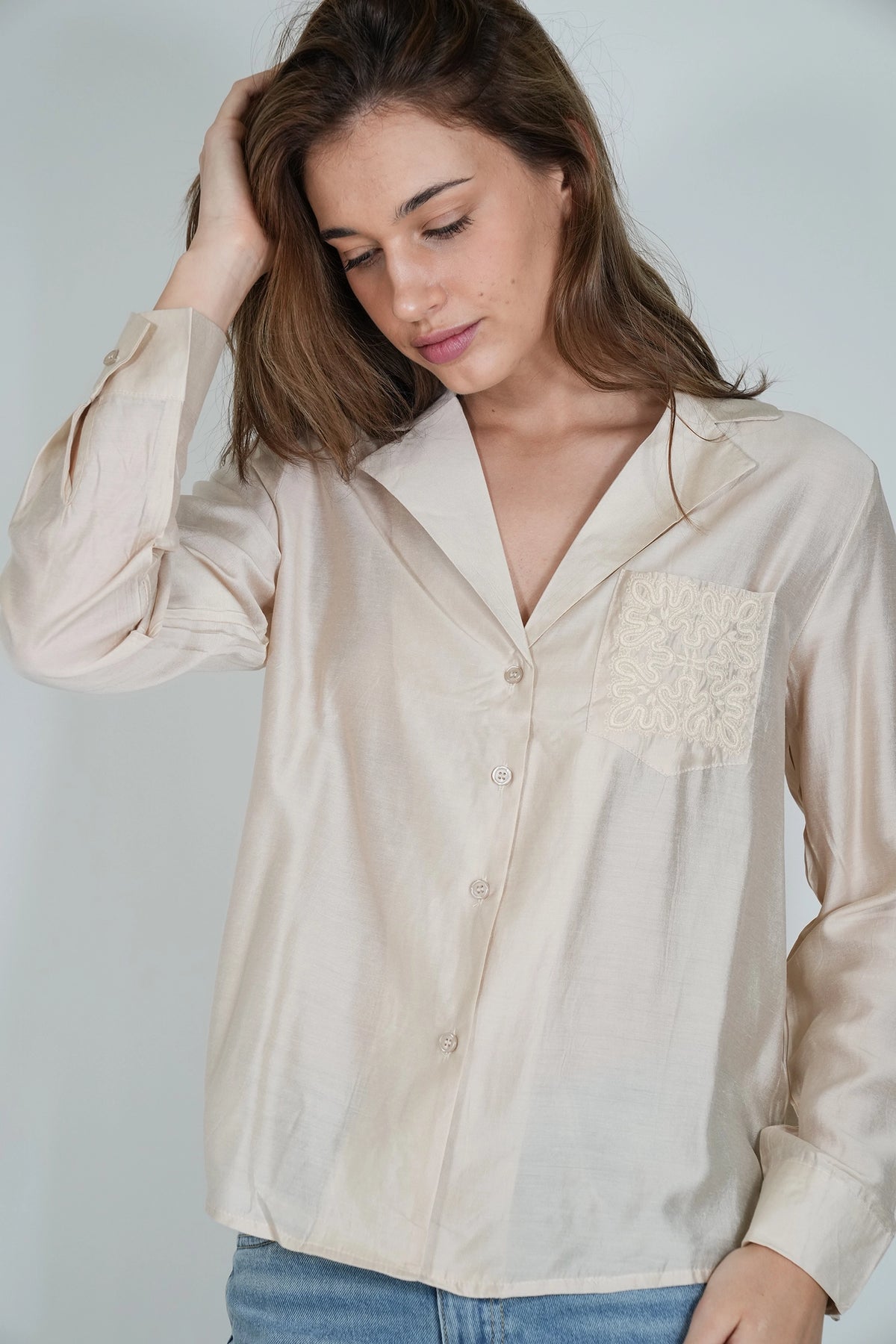 A woman wearing a light beige long-sleeve button-up shirt with a decorative embroidered pocket on the chest, paired with blue jeans, set against a plain light gray background.