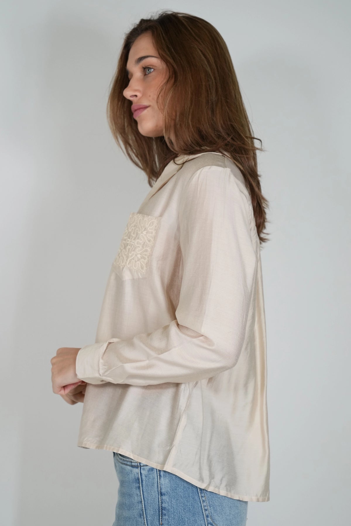 A woman wearing a light beige long-sleeve blouse with a textured lace chest pocket, paired with blue jeans, shown from a side profile against a plain light gray background.