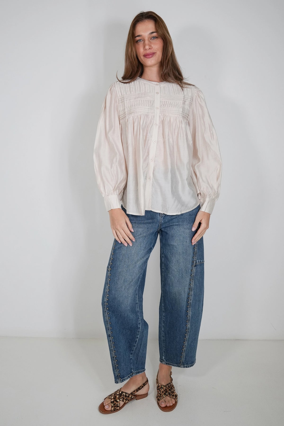 A woman modeling a cream-colored long-sleeve blouse with smocked detailing on the chest, paired with wide-leg blue jeans and leopard-print flat sandals, standing against a plain light gray studio background.