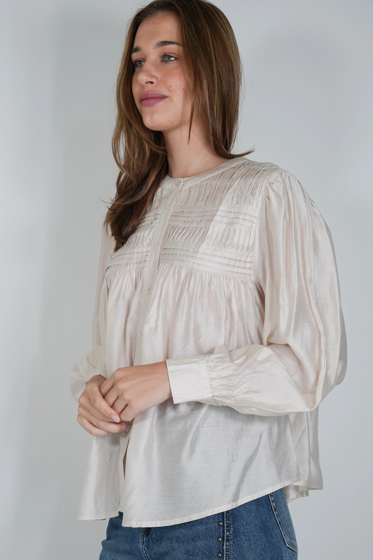 A woman wearing a cream-colored long-sleeve blouse with gathered smocked detailing on the chest and cuffs, paired with blue jeans, photographed against a neutral light gray background.