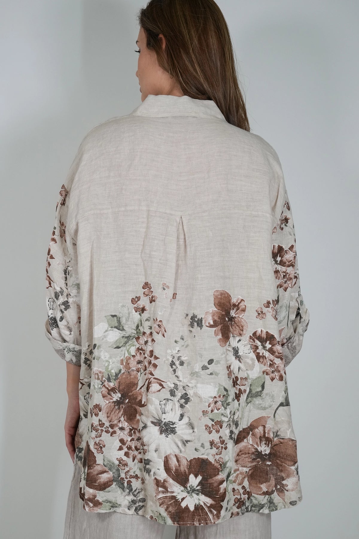 A woman viewed from behind wearing a light beige linen shirt with a brown and muted green floral print concentrated along the lower back and sleeves, set against a plain light gray background.