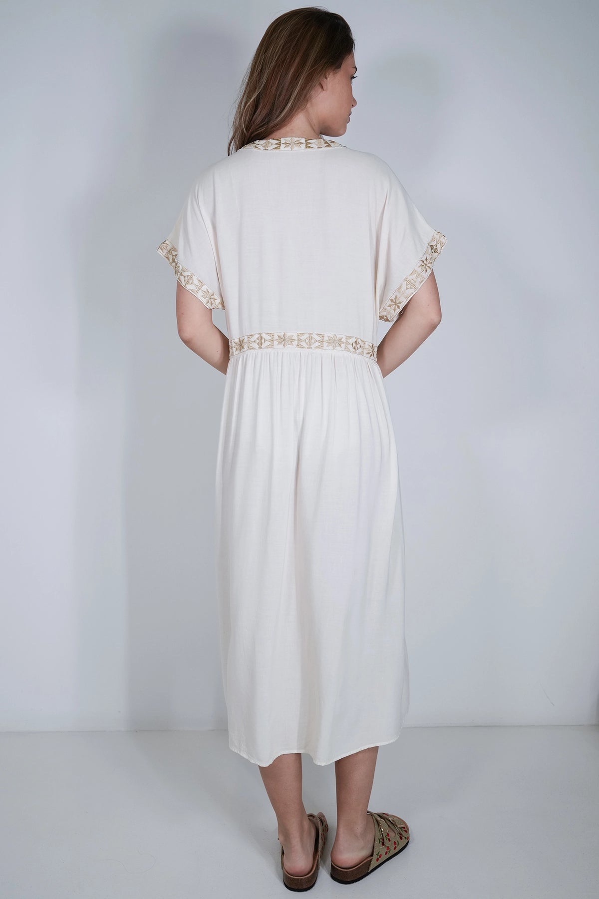 A woman viewed from the back wearing a cream-colored midi dress with gold lace trim on the collar and sleeve cuffs, paired with brown flat sandals, set against a plain light gray studio background.