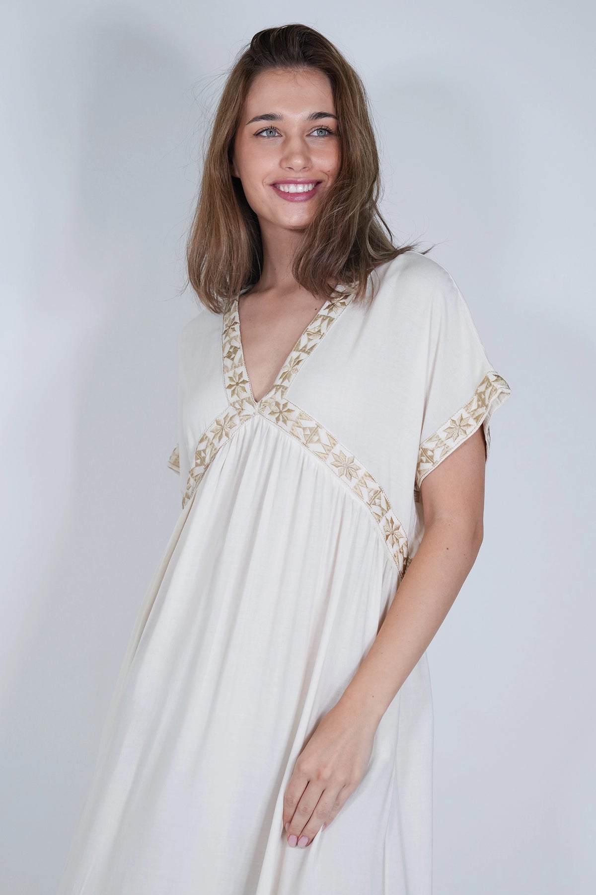 A woman wearing a cream-colored kaftan dress with gold floral embroidery along the V-neckline and sleeve cuffs, posing against a light gray studio background.