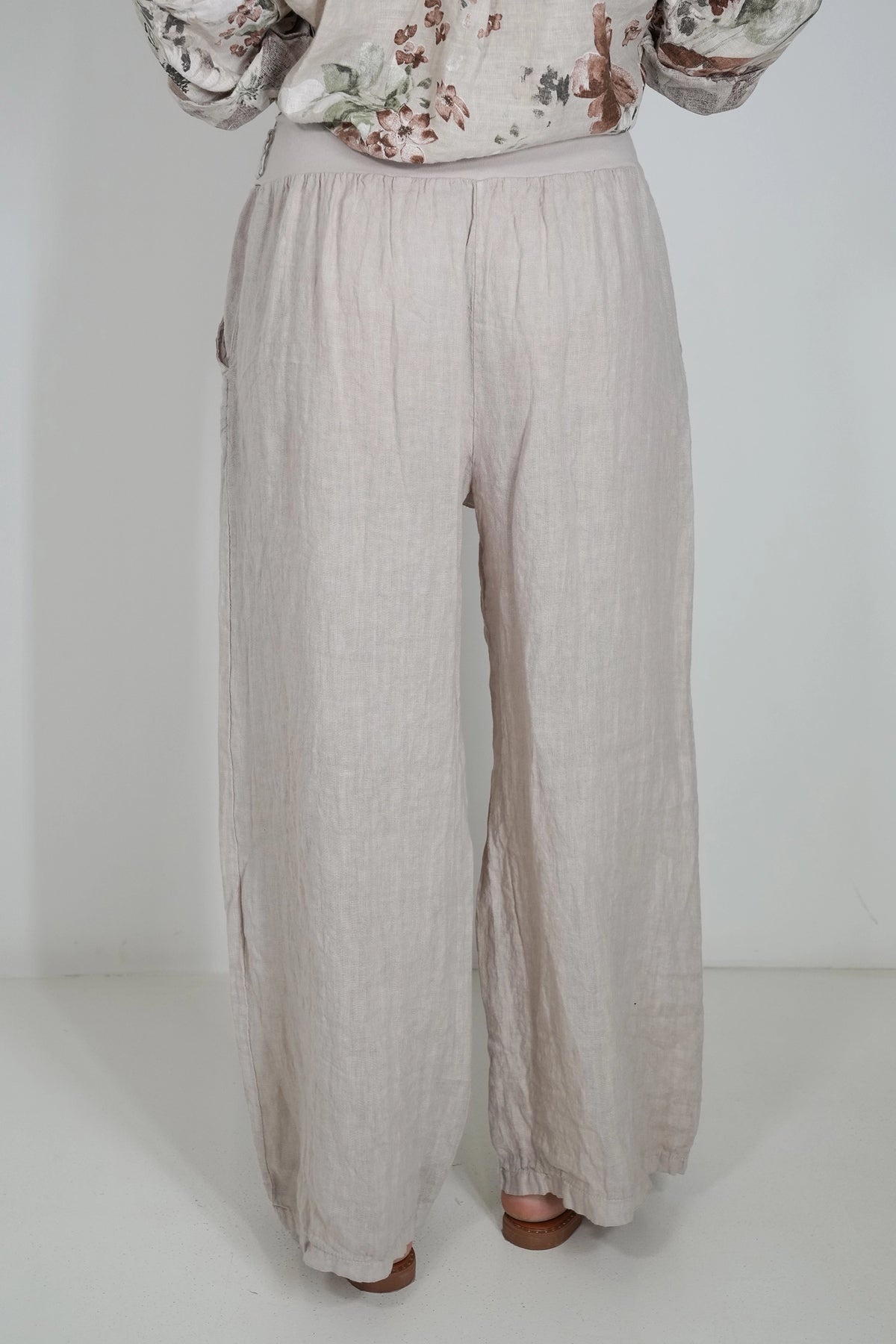 A person wearing loose, wide-leg linen pants in a light beige or oatmeal color, paired with a floral long-sleeve top, shown from the waist down against a plain white studio background.