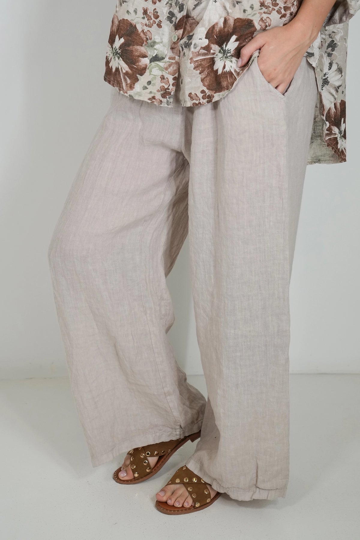 A person wearing loose, wide-leg linen pants in a light beige or oatmeal color, paired with brown strappy sandals and a floral top, standing against a plain white studio background.
