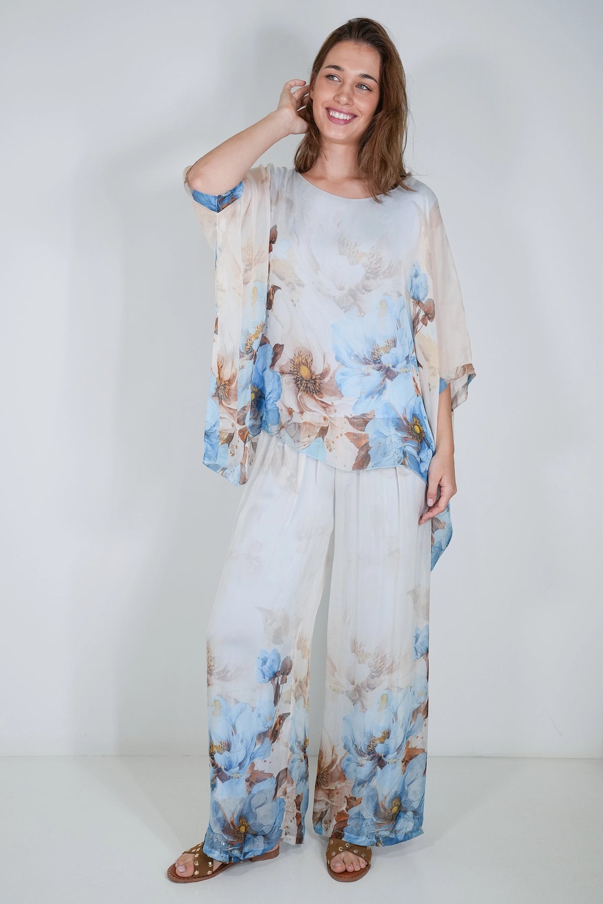 A woman modeling a matching two-piece sheer lounge set with a blue and brown floral print on a white base, featuring a loose short-sleeve top and wide-leg pants, set against a plain white studio background.