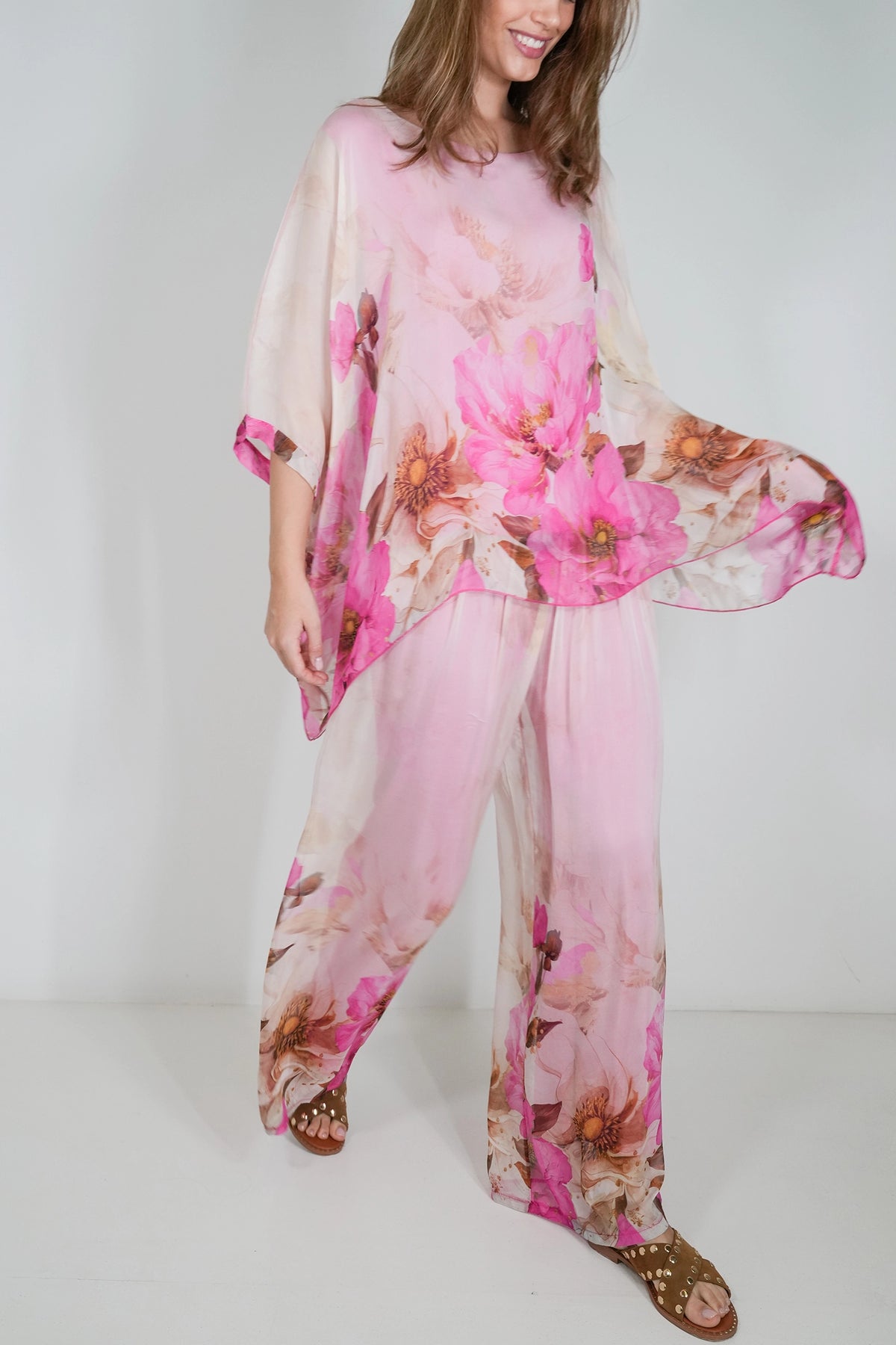 A woman wearing a light pink sheer two-piece outfit with a large pink floral print, consisting of a loose short-sleeve top and matching wide-leg pants, paired with brown strappy sandals against a plain white studio background.