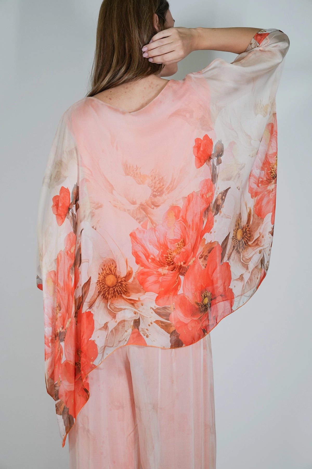 A woman shown from the back wearing a sheer, flowing pink top with a large coral and red floral print, paired with matching sheer pink pants, against a light gray studio background.