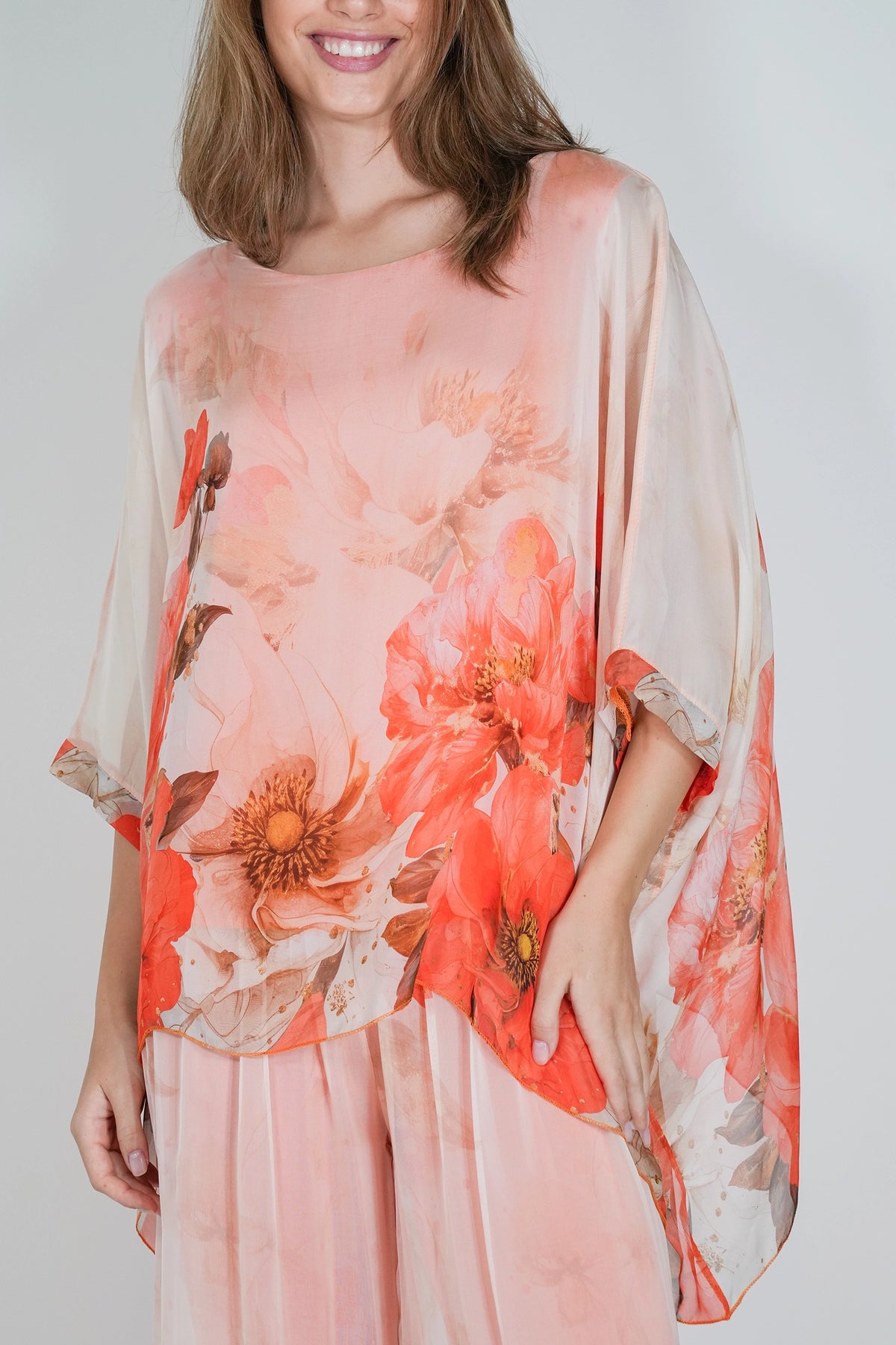 A woman wearing a loose, semi-sheer peach and coral floral top with wide draped sleeves, paired with matching pink bottoms, against a light gray studio background.