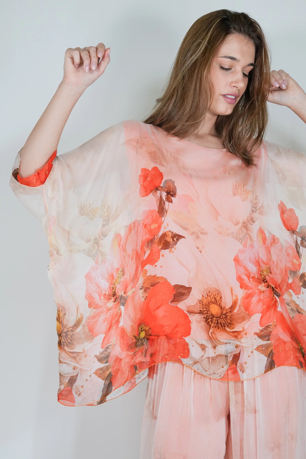 A woman wearing a sheer peach and coral floral kaftan top with wide draped sleeves, paired with matching pink bottoms, posed against a light gray studio background.