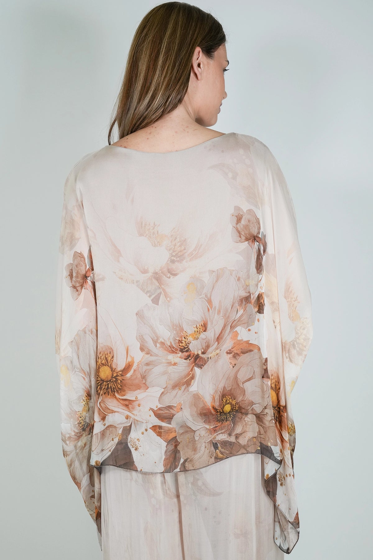 A woman shown from the back wearing a sheer, long-sleeve top with a large floral print in soft beige, brown, and muted orange tones, paired with light beige bottoms, against a plain light gray background.