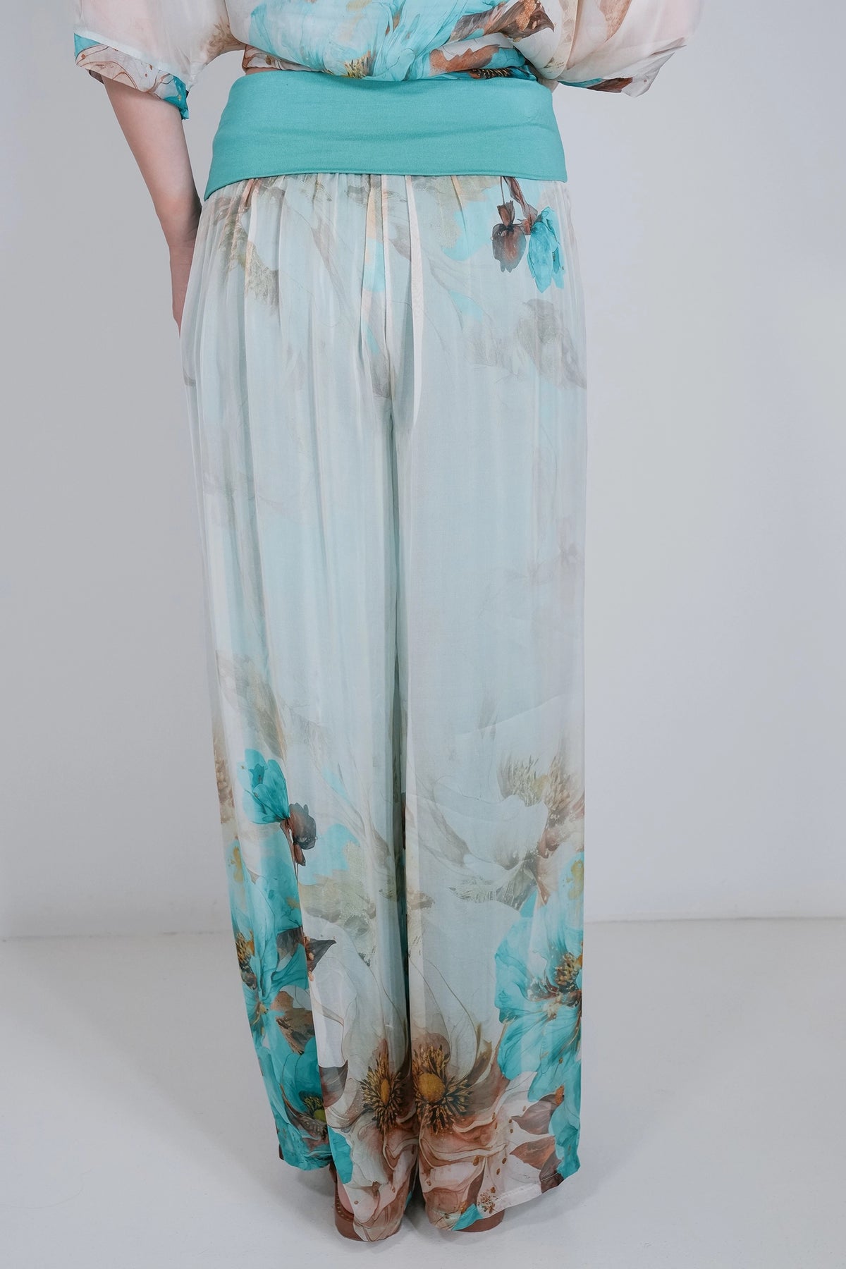 A person wearing wide-leg trousers with a light blue and white floral print, a turquoise waistband, and a matching turquoise top, shown from the back against a plain light gray studio background.