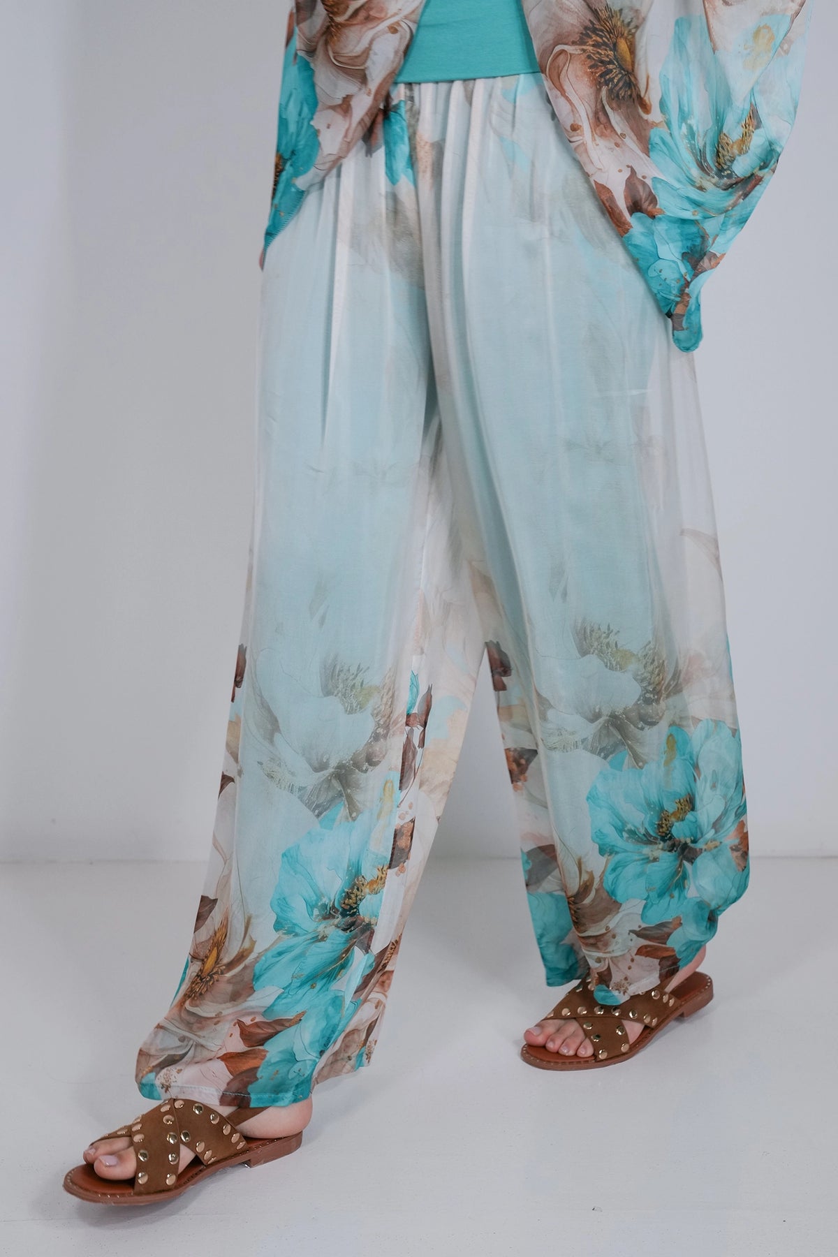 A person wearing loose, wide-leg sheer pants with a large floral print in turquoise, beige, and brown, paired with brown studded sandals against a plain white studio background.