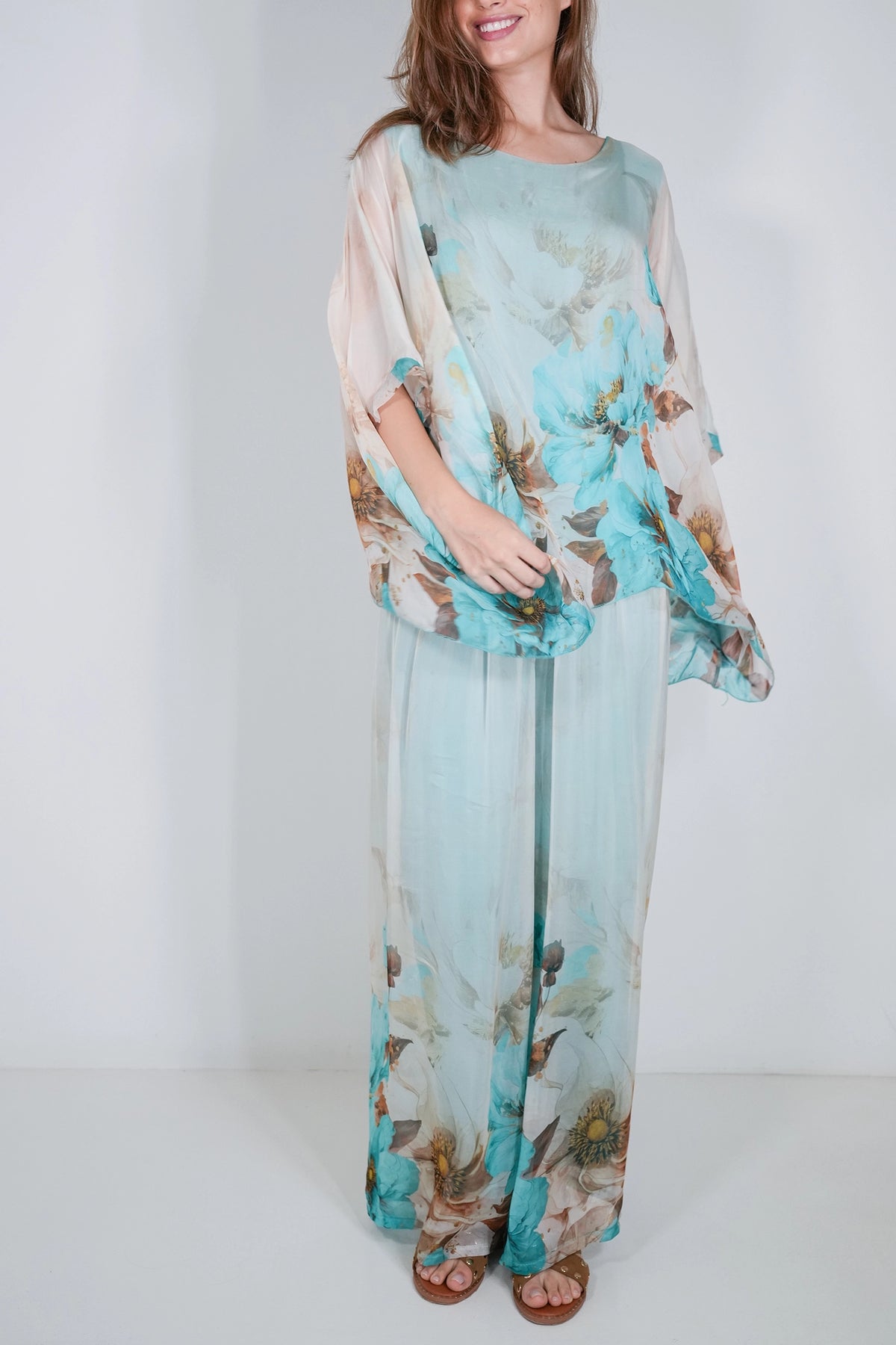 A woman modeling a light blue and white floral printed two-piece outfit with sheer draped sleeves and wide-leg pants, standing against a plain white studio background.